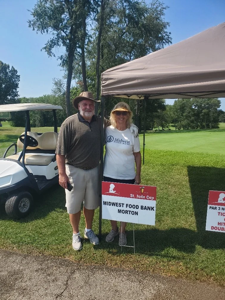 A man and woman standing under a canopy at a golf course, smiling for a photo. The woman is wearing a white shirt with a logo for Midwest Food Bank. There are signs indicating a golf event for Midwest Food Bank Morton and a golf cart nearby.