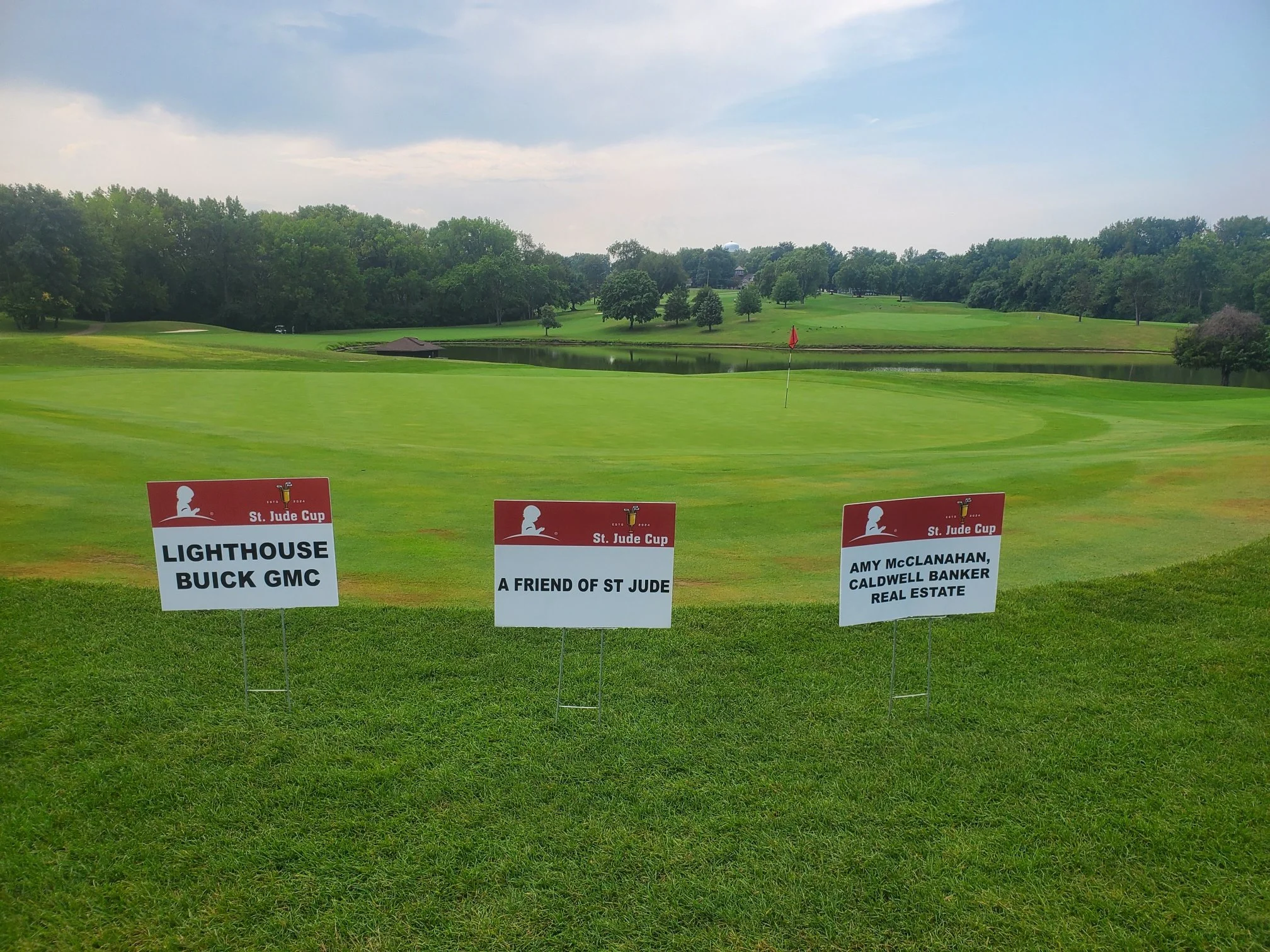Three signs on a golf course with a green, water hazard, trees, and a cloudy sky in the background.