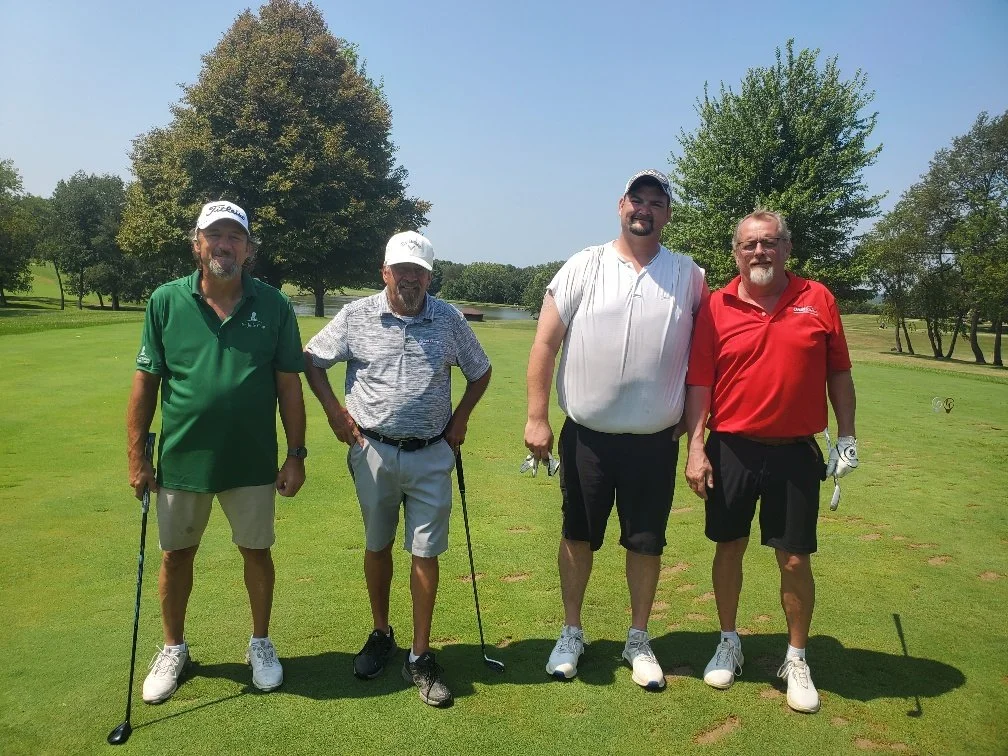 Group of four men standing on a golf course holding golf clubs, with trees and a pond in the background on a sunny day.