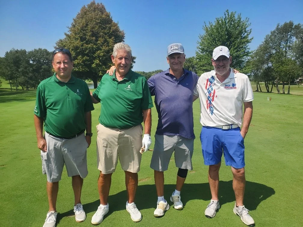 Four men standing on a golf course, smiling, with trees and blue sky in the background.