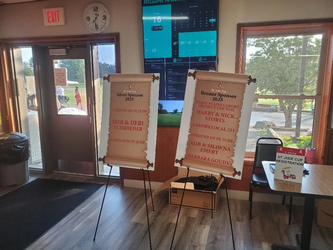 Indoor registration area for the St. Jude Cup with two standing banners displaying sponsors and recipients, a table with a sign-in sheet, a digital screen showing weather and event information, an exit door, and a window showing the outside view of t