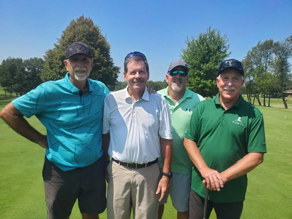 Four men standing together on a golf course, smiling, with trees and clear blue sky in the background.