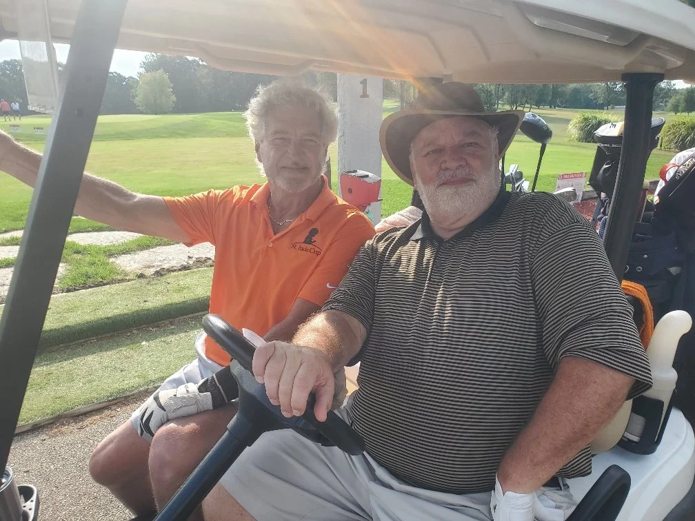 Two older men sitting in a golf cart on a golf course, with one man holding the steering wheel and the other sitting beside him, both smiling at the camera.