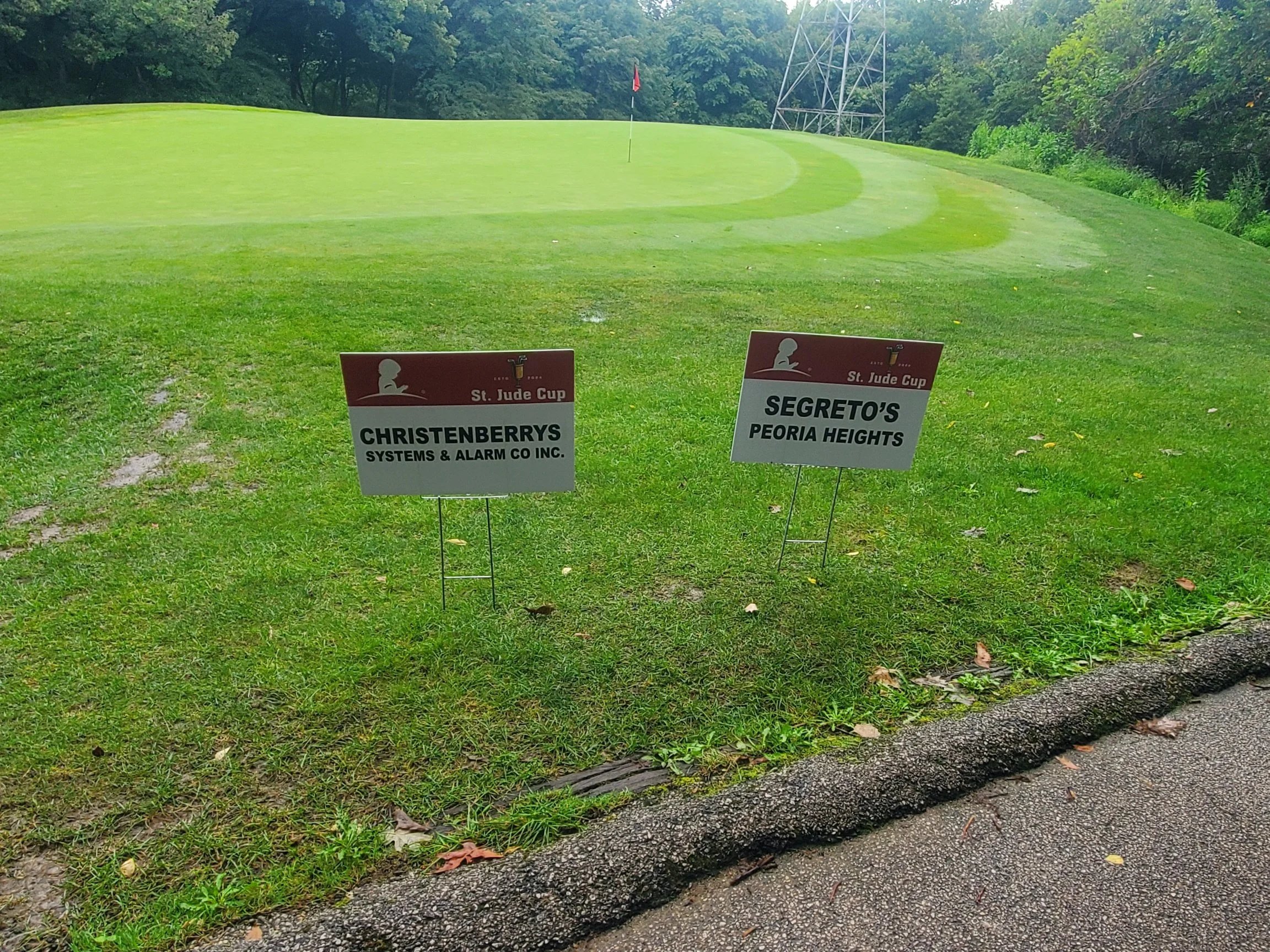Golf course green with two signs in the foreground, one for Christenberrys and one for Segreto's, with a flag on the green and trees in the background.