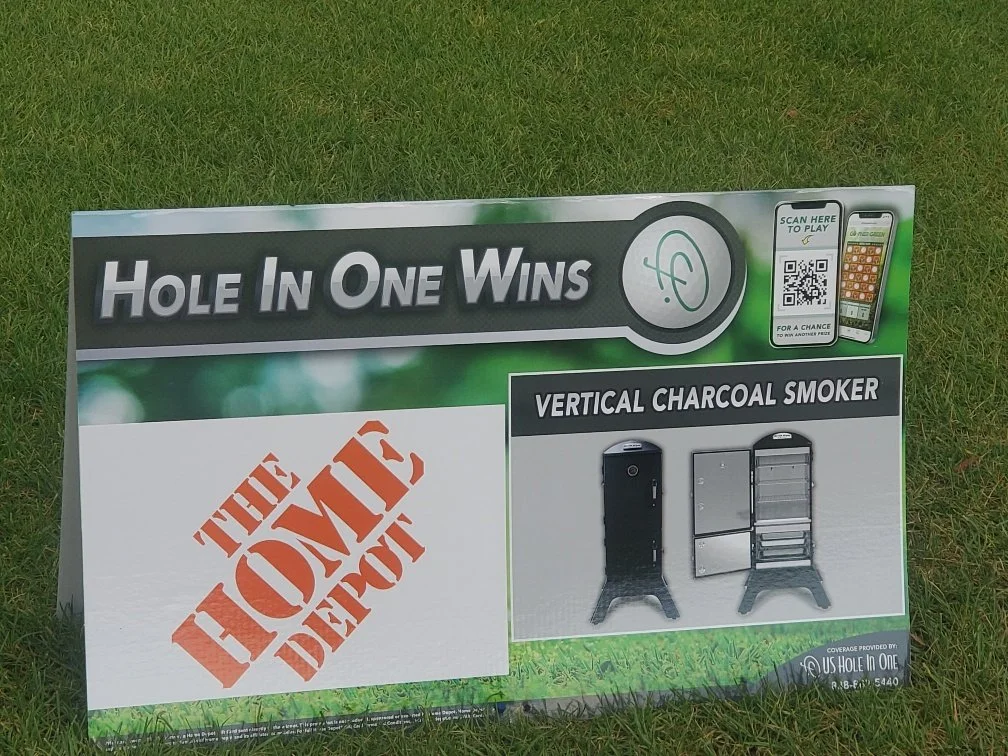 Sign for a game called 'Hole in One Wins' with a section titled 'The Home Depot', a QR code for a mobile game, and a depiction of a vertical charcoal smoker with two views, on a grassy area.