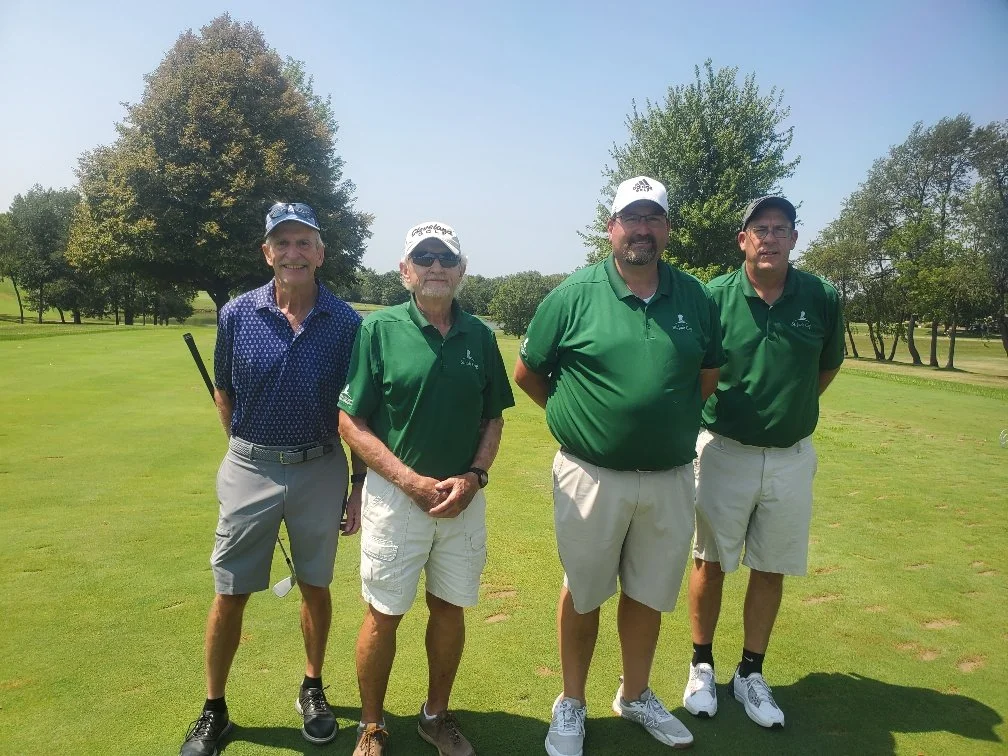Four men standing on a golf course, smiling, with trees in the background. They are wearing golf attire, with three in green polo shirts and one in a blue patterned shirt.