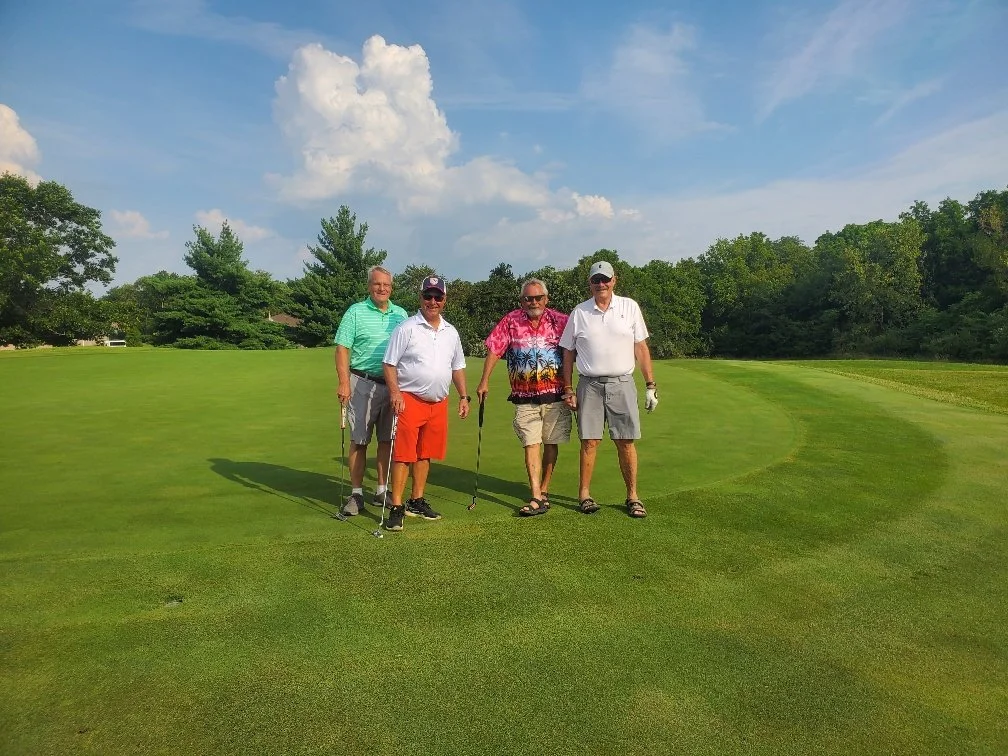 Four men standing on a golf course, holding golf clubs, with trees, grass, and a partly cloudy blue sky in the background.