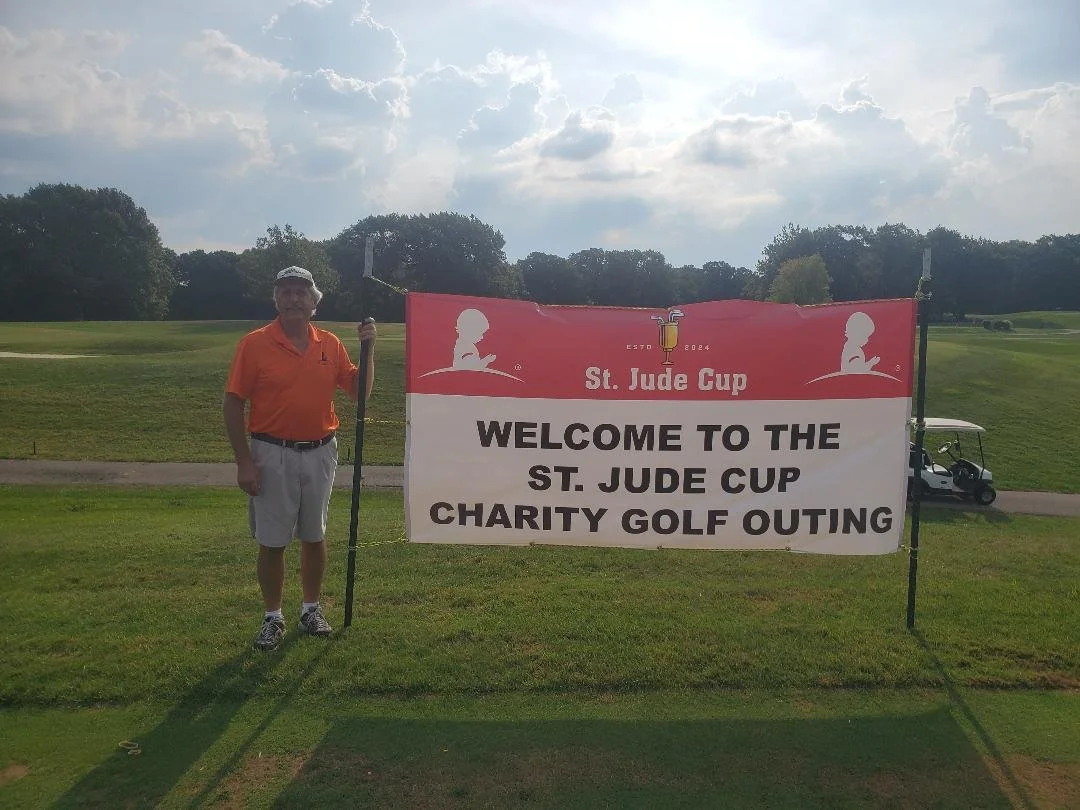 A man in an orange shirt and white shorts holding a golf club standing next to a large sign on a golf course. The sign reads 'St. Jude Cup Welcome to the St. Jude Cup Charity Golf Outing' with logos and decorative elements.