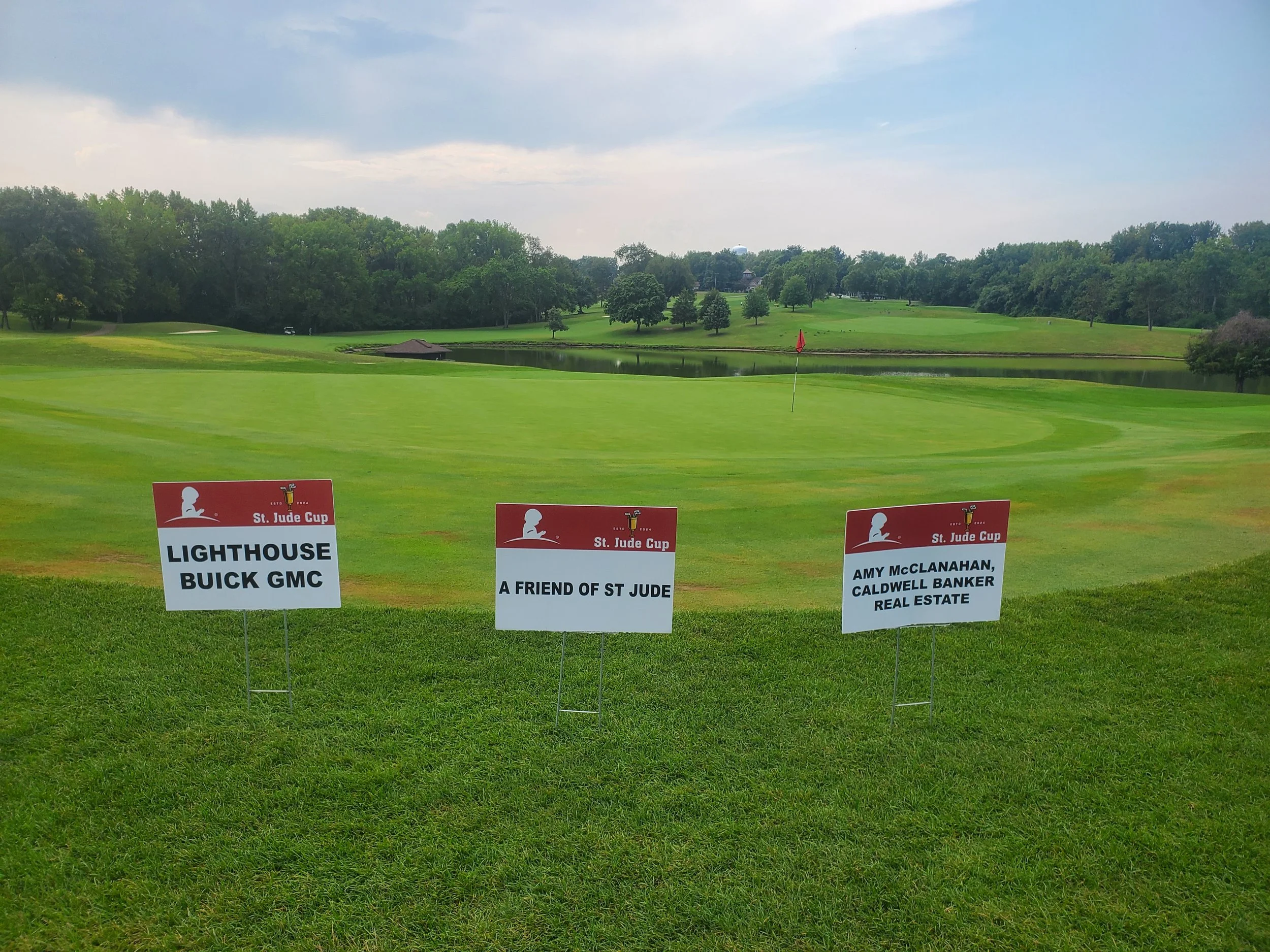 Golf course with three sponsor signs on the grass in the foreground, a pond, trees, and cloudy sky in the background.