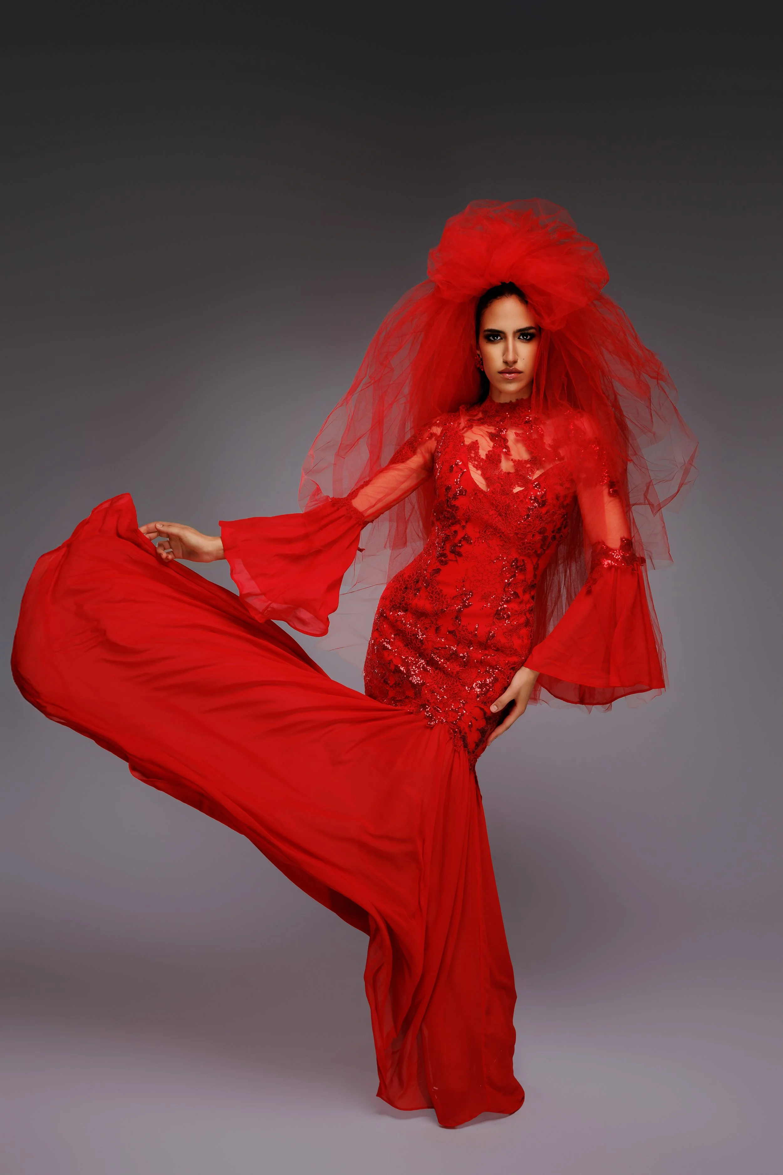 Femme portant une robe de soirée rouge avec un voile assorti, posant contre un fond gris.