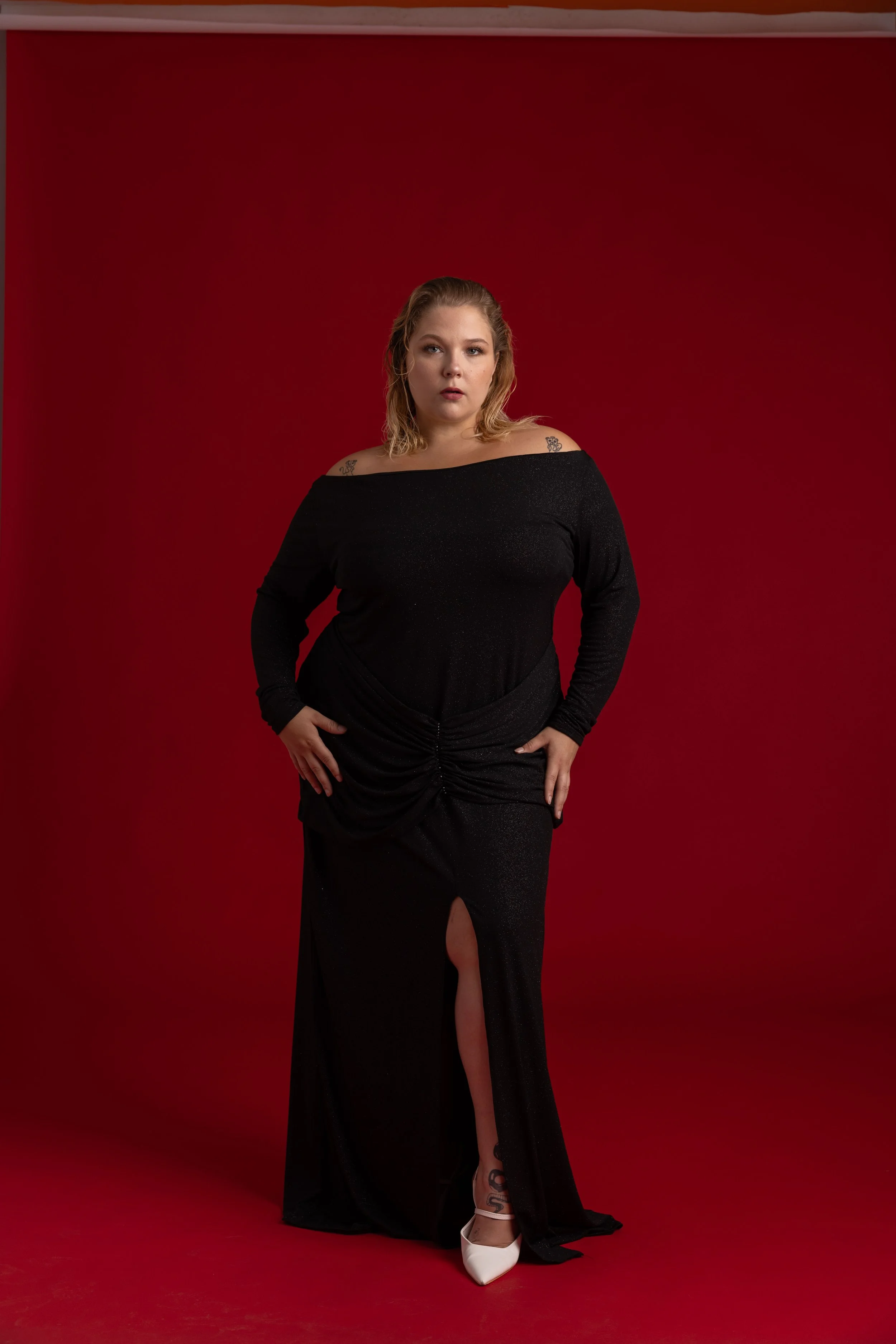Femme debout en robe noire longue à décolleté plongé, avec un fond rouge.