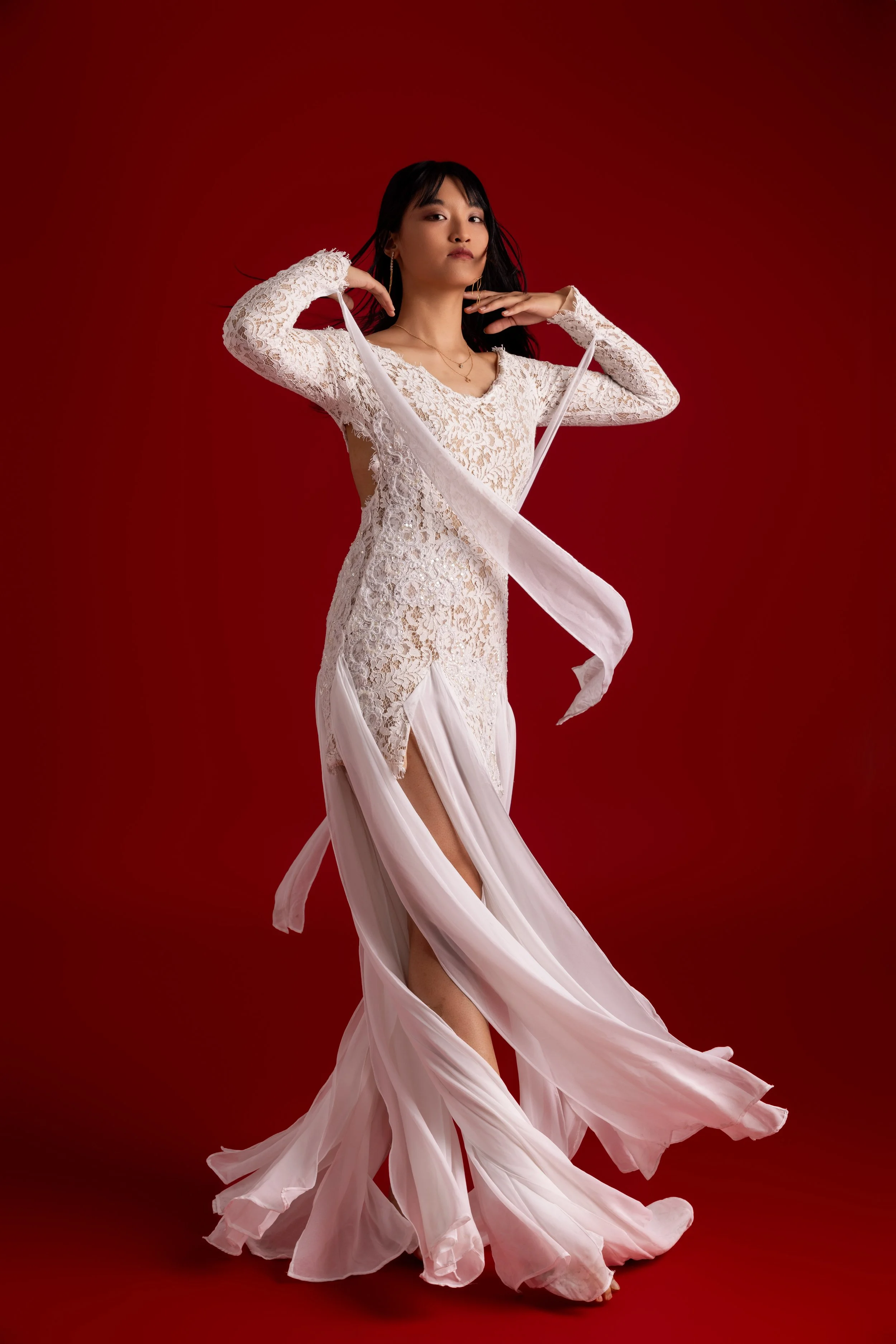 Une femme en robe longue en dentelle blanche et tissu léger, posant devant un fond rouge.