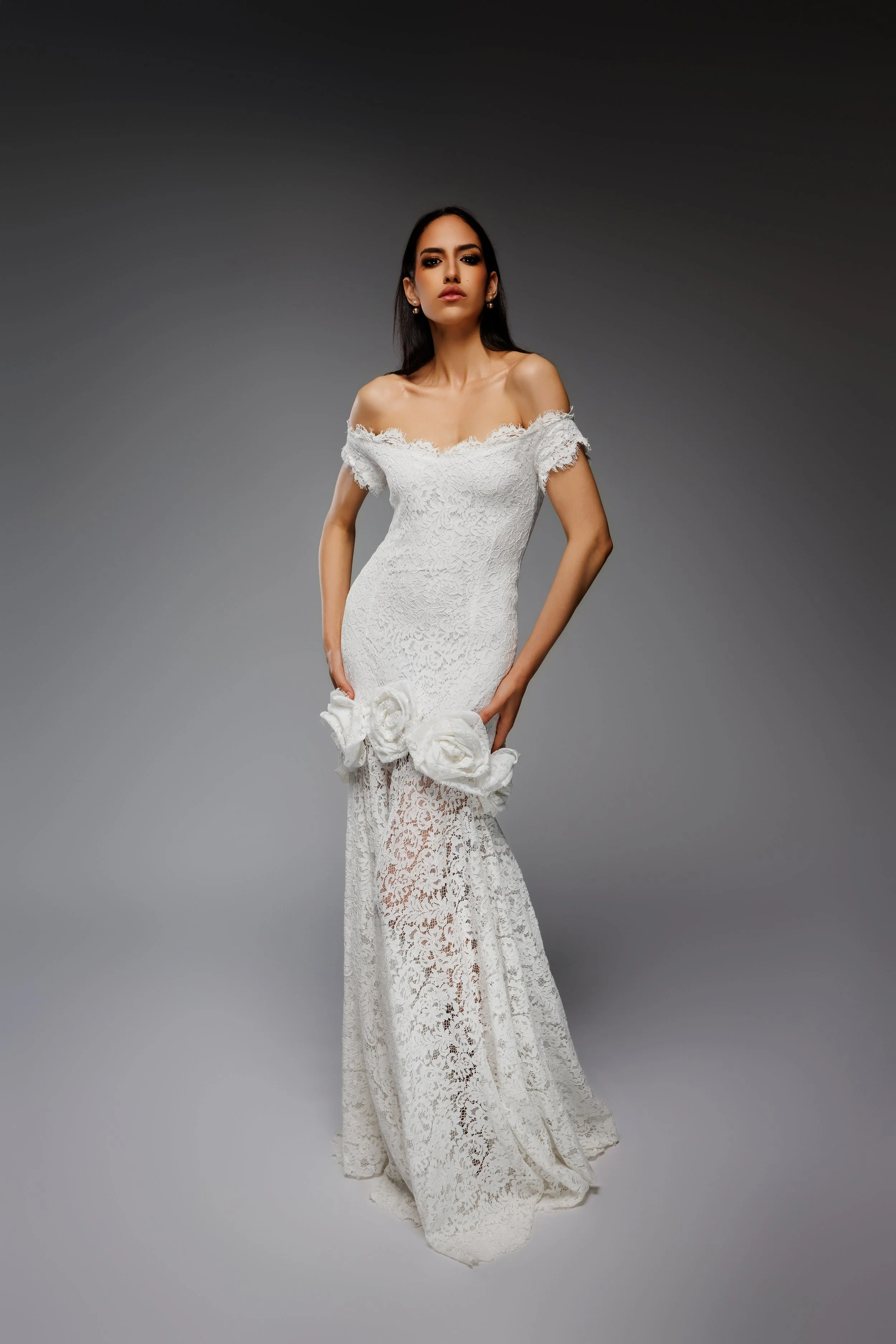 Une femme porte une robe de mariage blanche en dentelle avec des détails floraux et un décolleté en vue, posant devant un fond gris