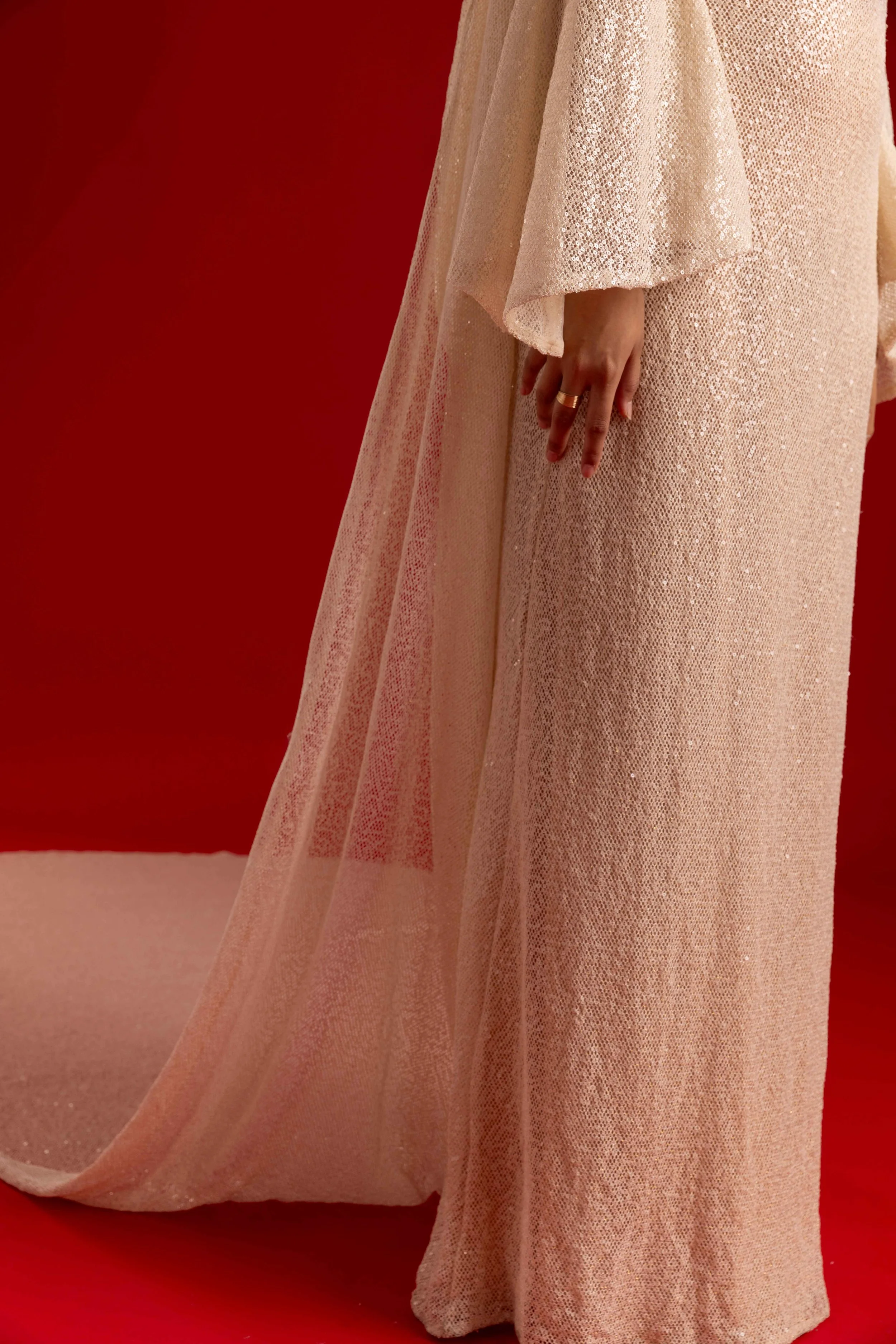 Une personne portant une robe longue beige avec des détails brillants, debout devant un fond rouge.