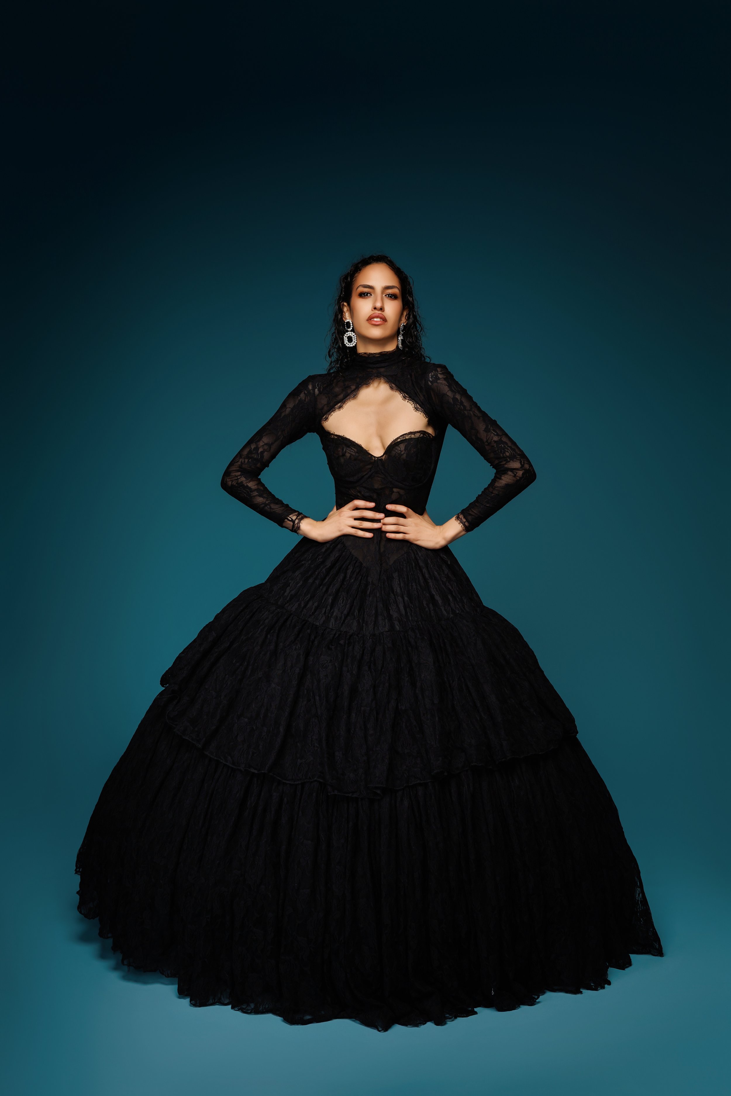 Une femme portant une robe longue noire en dentelle, avec des détails transparents, pose contre un fond dégradé de bleu.