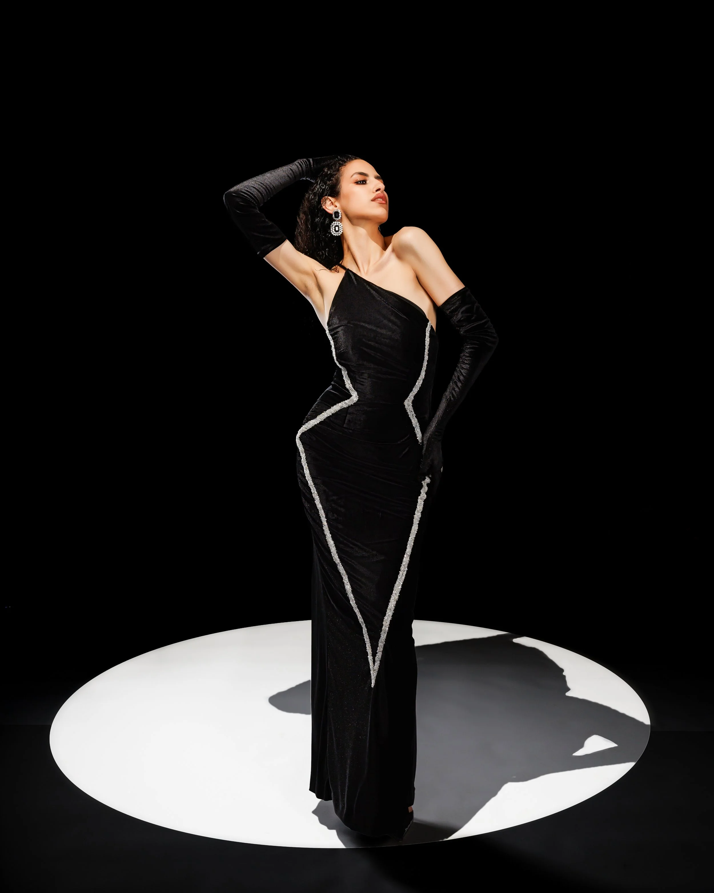 Femme élégante en robe noire avec dentelle argentée, portant de longues gants noirs, posant sur un fond noir avec un cercle blanc au sol.