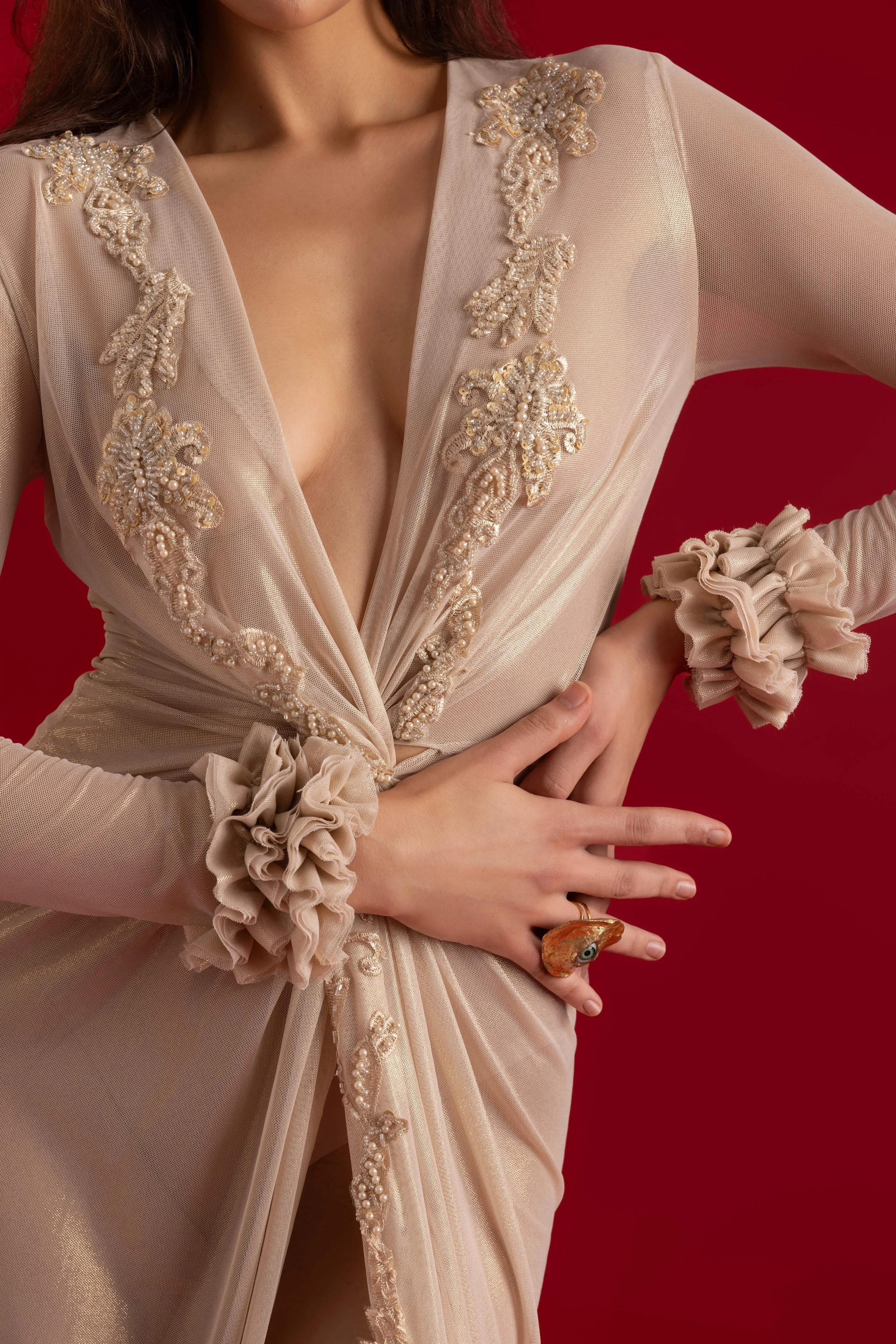 Femme portant une robe beige ornée de broderies de perles, avec des manches en tulle et des accessoires de cheveux en tissu. La femme porte également une bague fantaisie en forme d'animal.