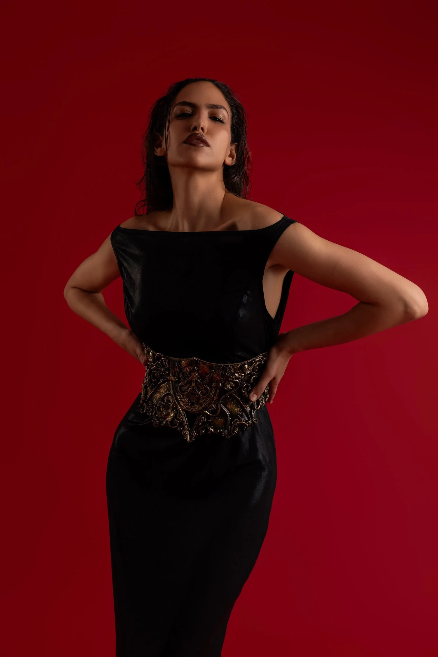 Une femme posant devant un fond rouge, portant une robe noire avec une ceinture ornementée.