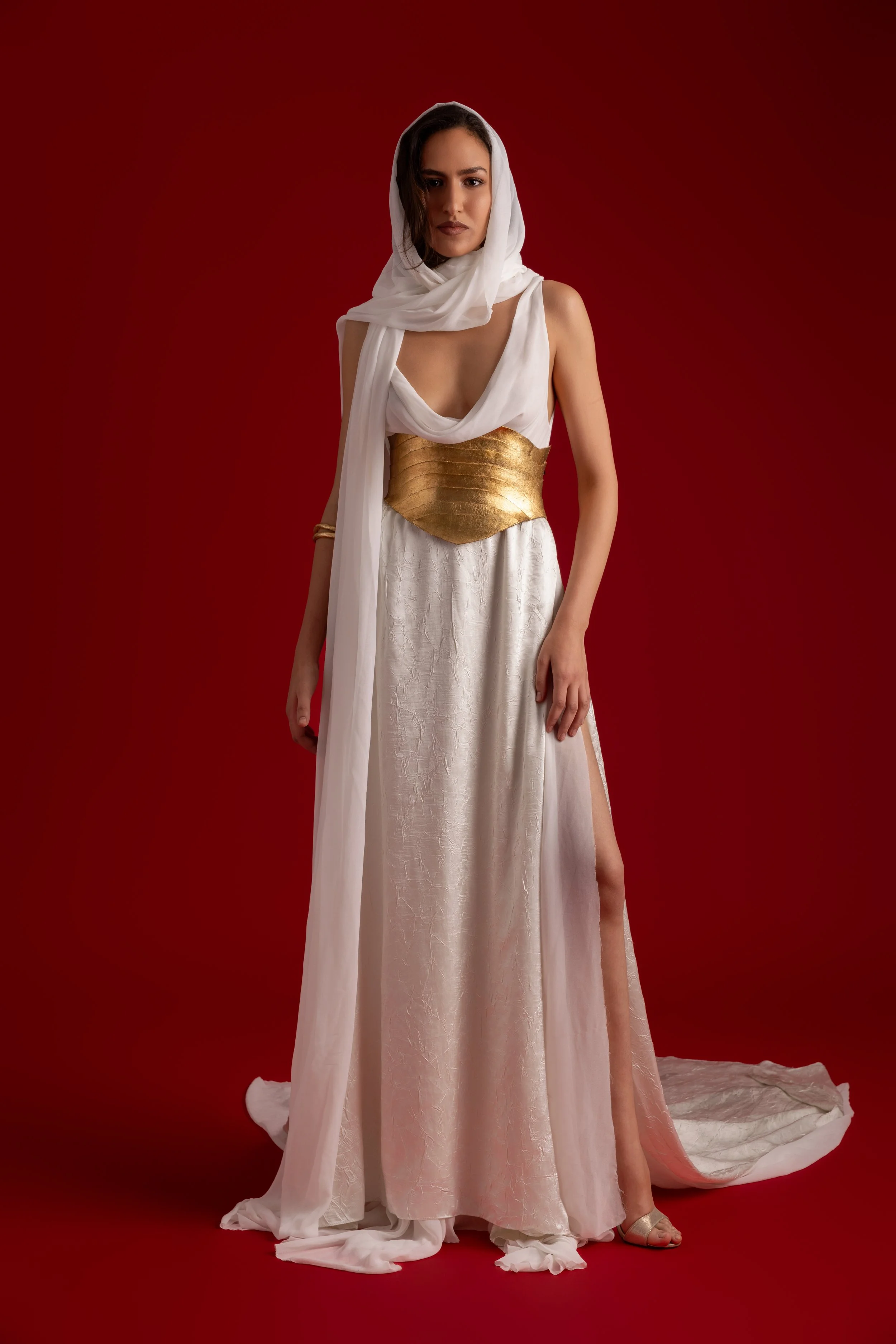 Femme portant une robe longue de style mythologique avec un châle blanc et une ceinture dorée, sur un fond rouge.