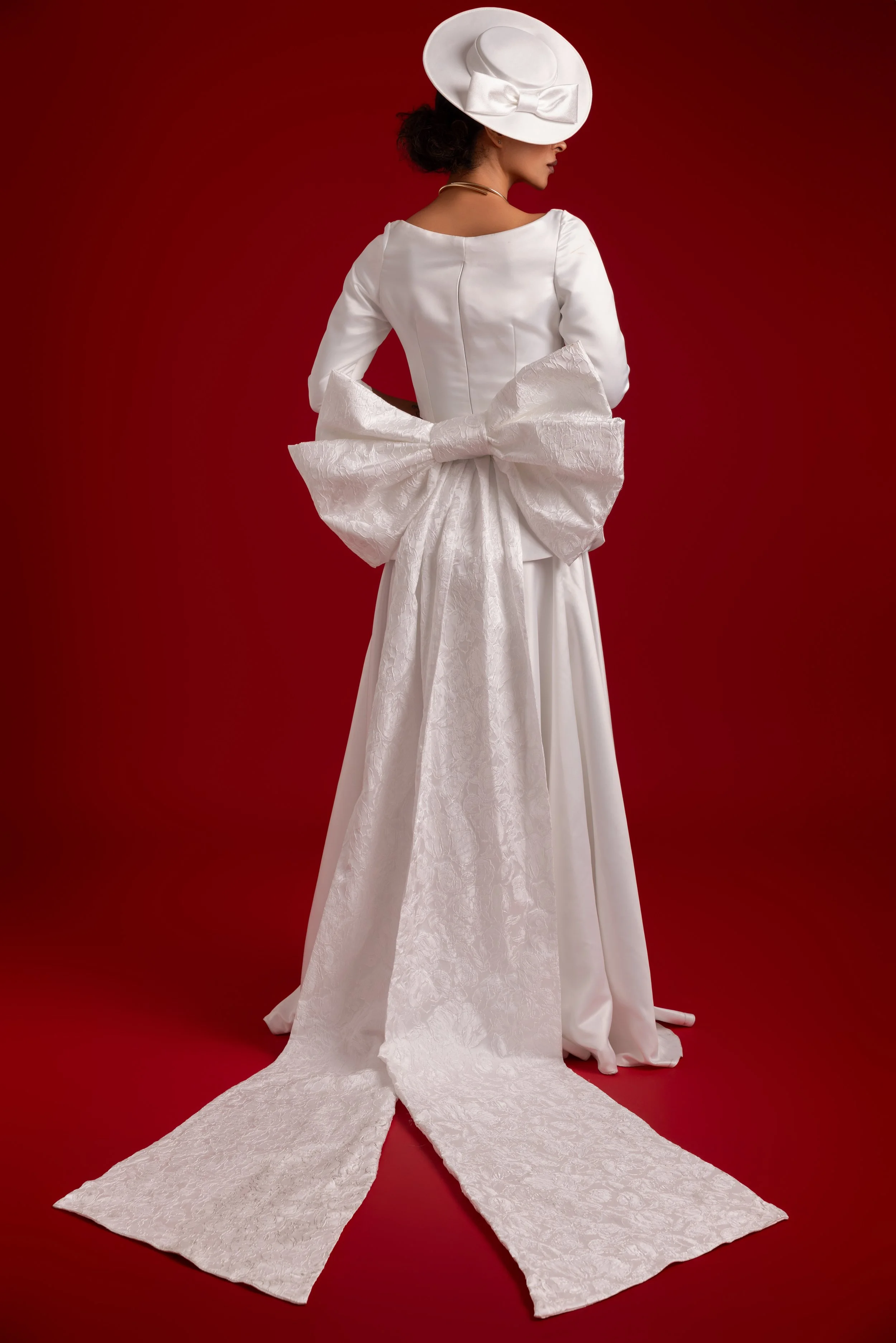 Femme en robe blanche élégante avec un grand ruban en tissu blanc dans le dos, portant un chapeau blanc avec un ruban, sur un fond rouge.