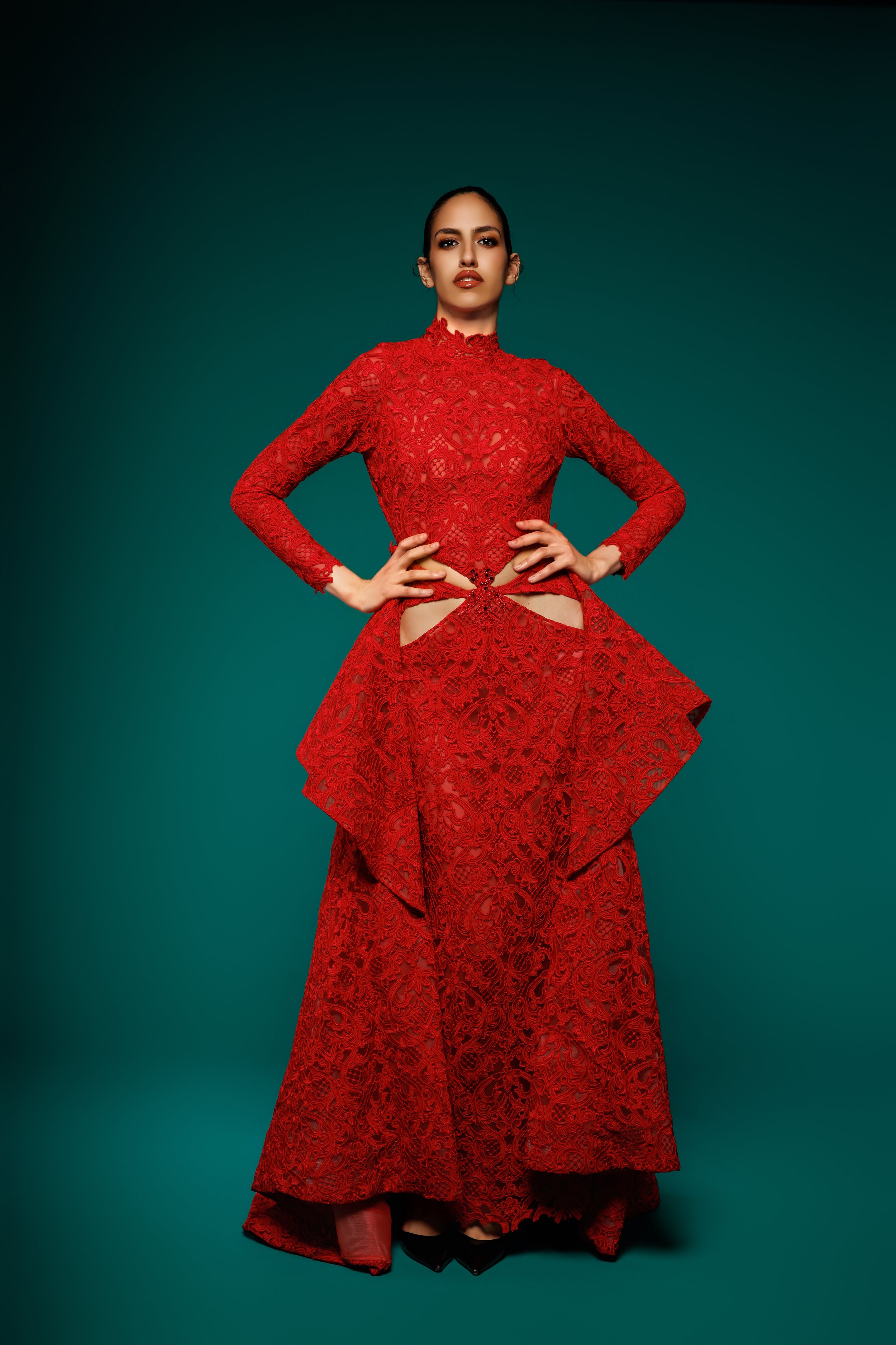 Femme en robe rouge en dentelle avec des découpes, posant avec les mains sur les hanches, contre un fond vert