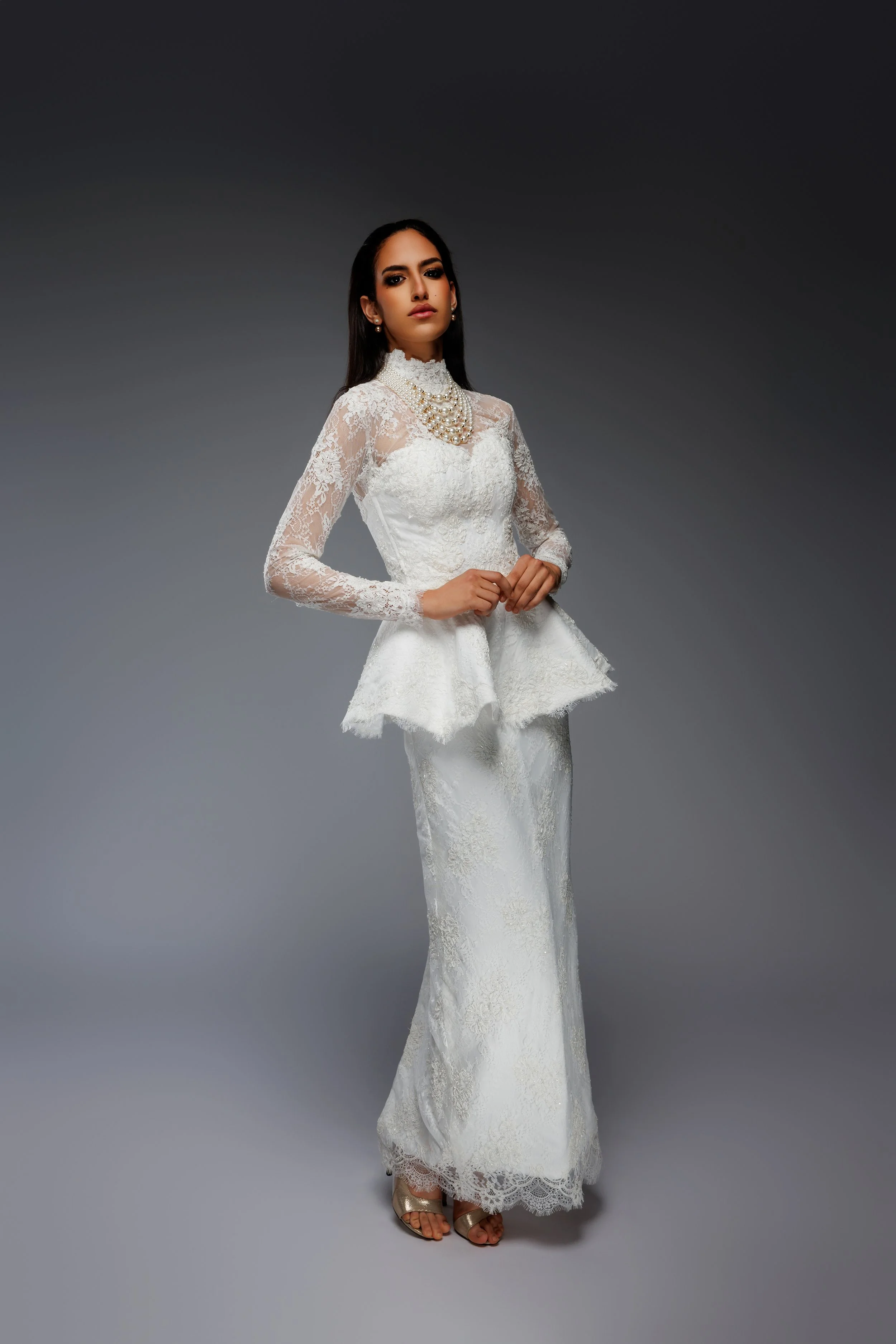 Femme portant une robe de mariage blanche en dentelle, avec des bijoux de perles, posant contre un fond gris.