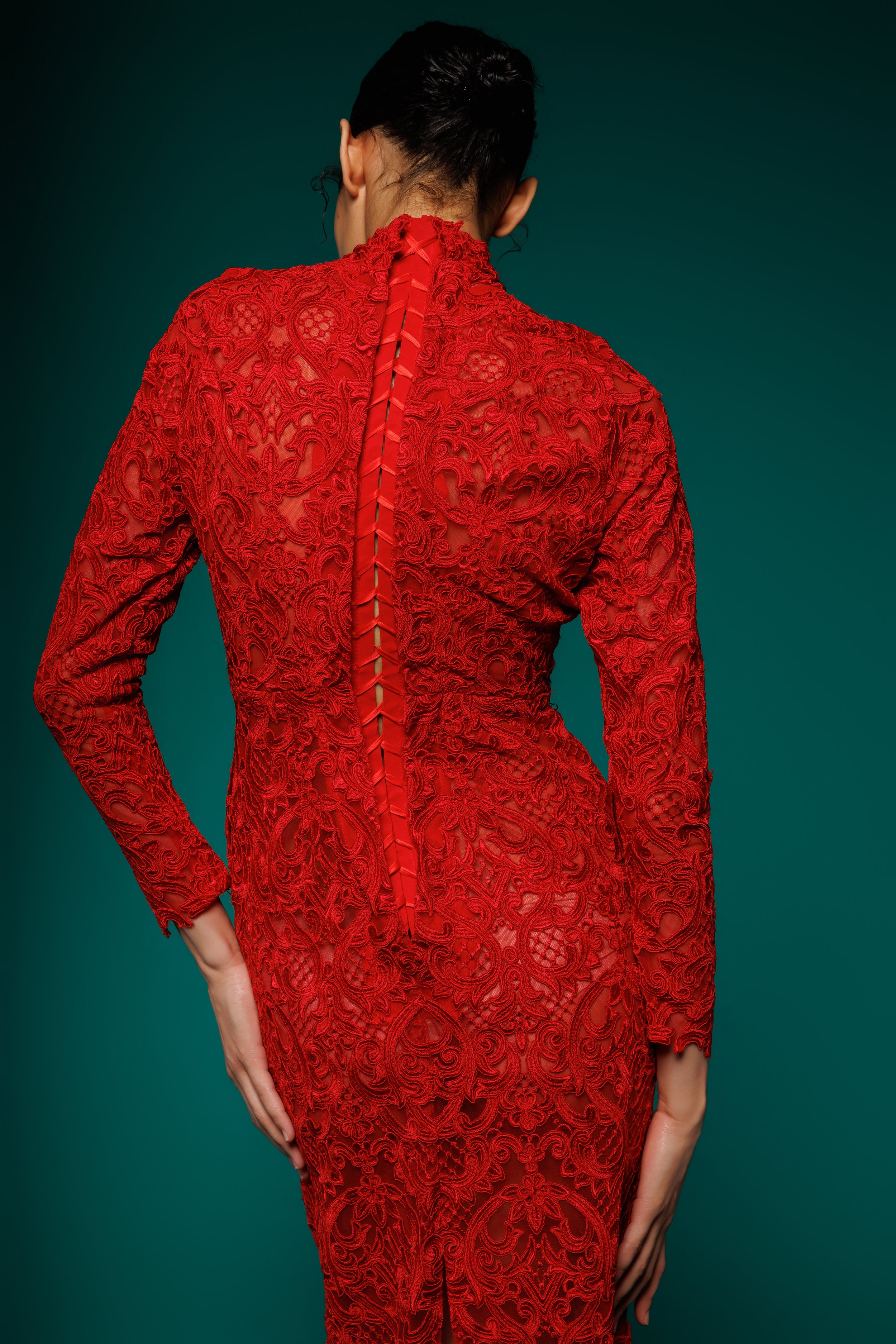 Femme de dos portant une robe longue en dentelle rouge avec un dos ouvert, contre un fond vert foncé.
