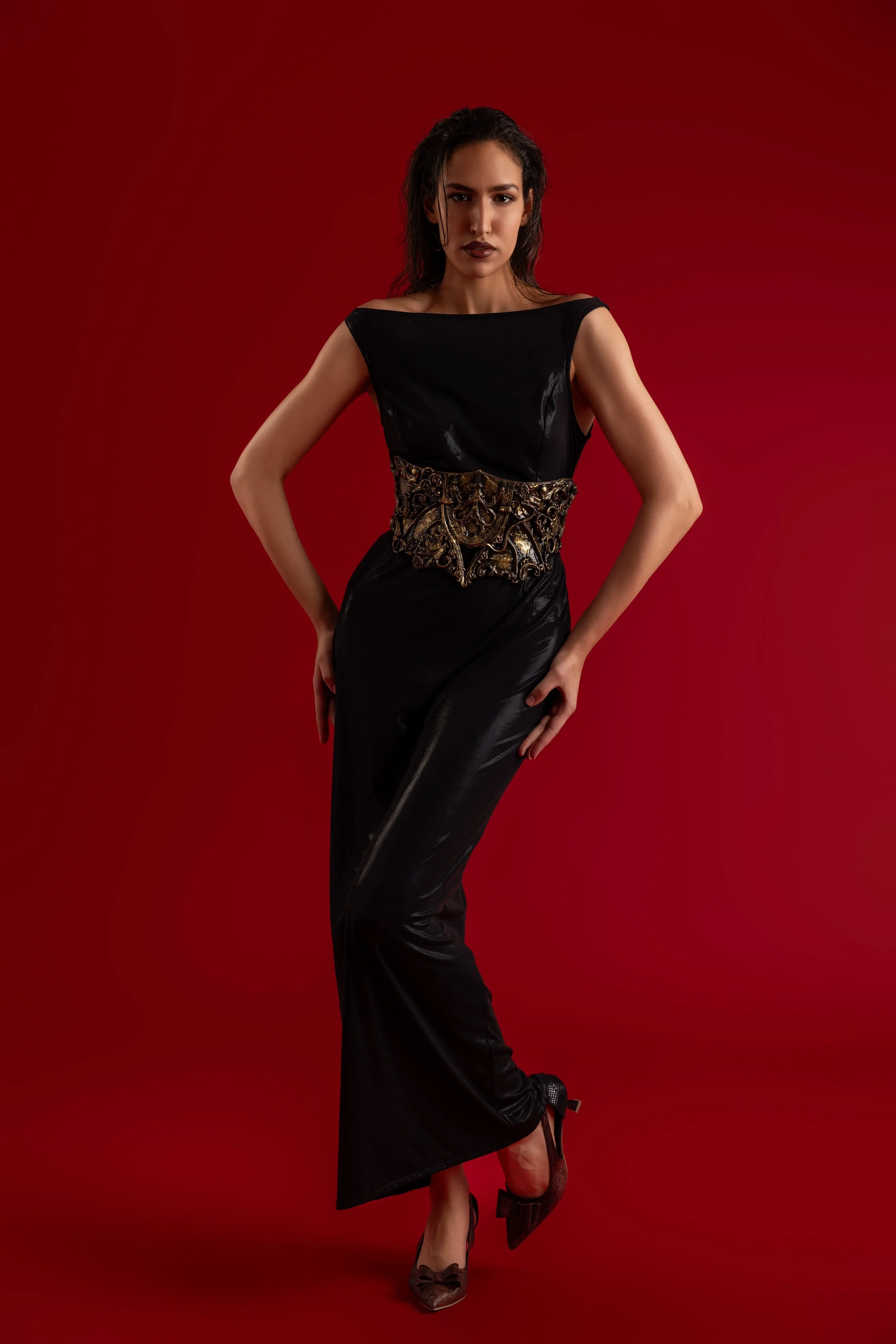 Une femme en robe noire élégante avec une ceinture dorée sculptée, posant contre un fond rouge.
