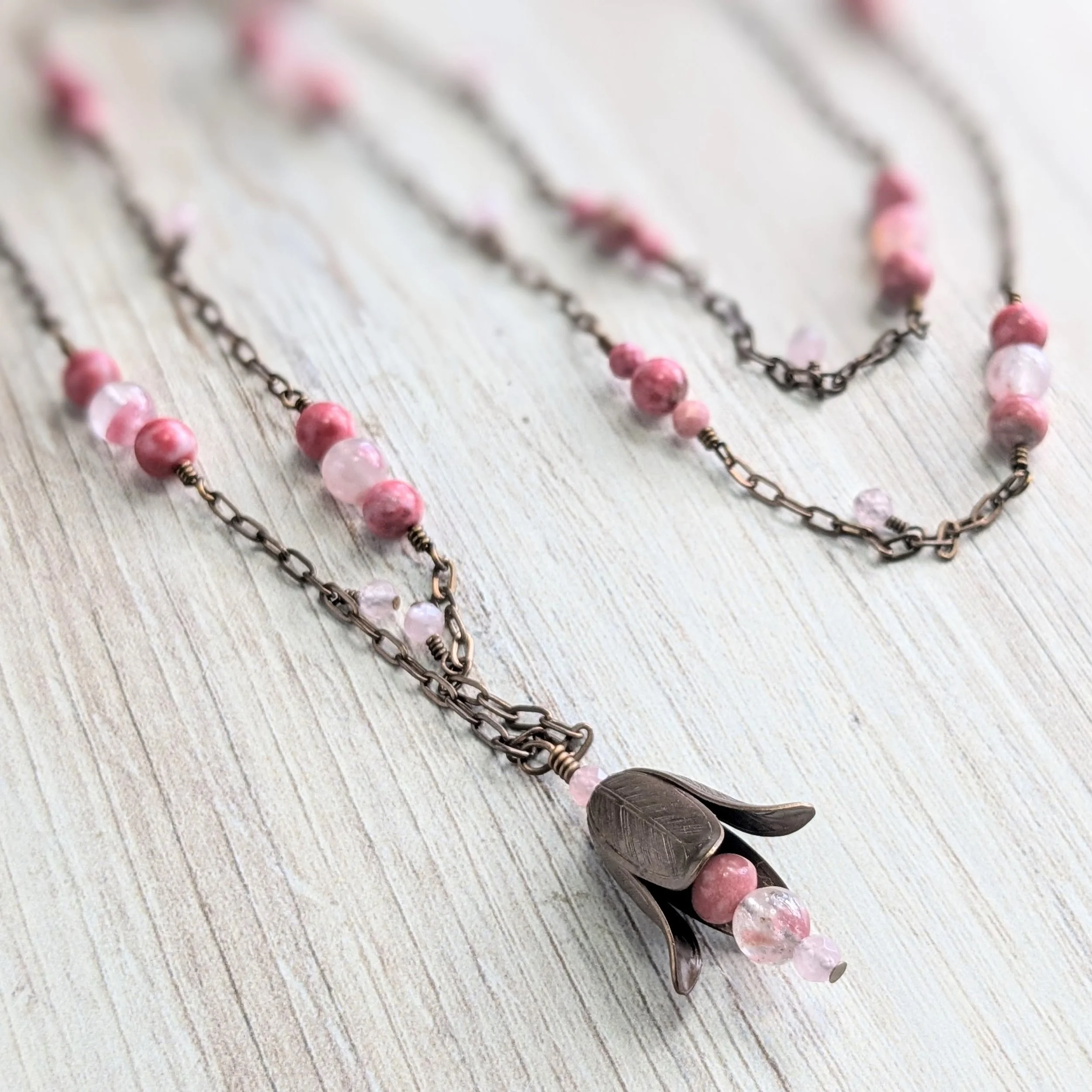 Necklace with pink beads, a dark metal chain, and a feather-shaped pendant with pink beads hanging from it, lying on a light wooden surface.