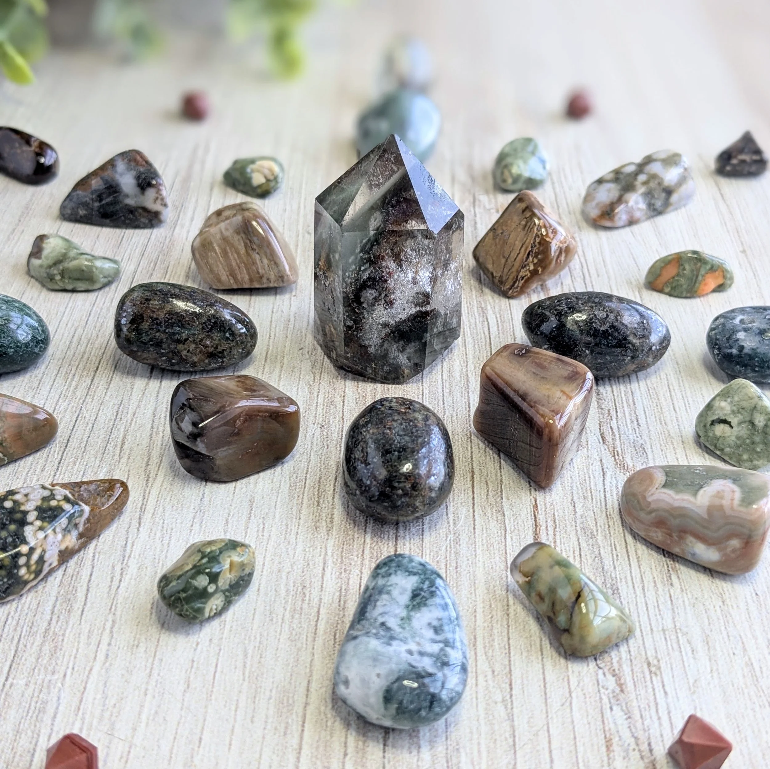 A collection of polished and raw stones and crystals, surrounding a Garden Quartz tower, arranged on a light wooden surface.
