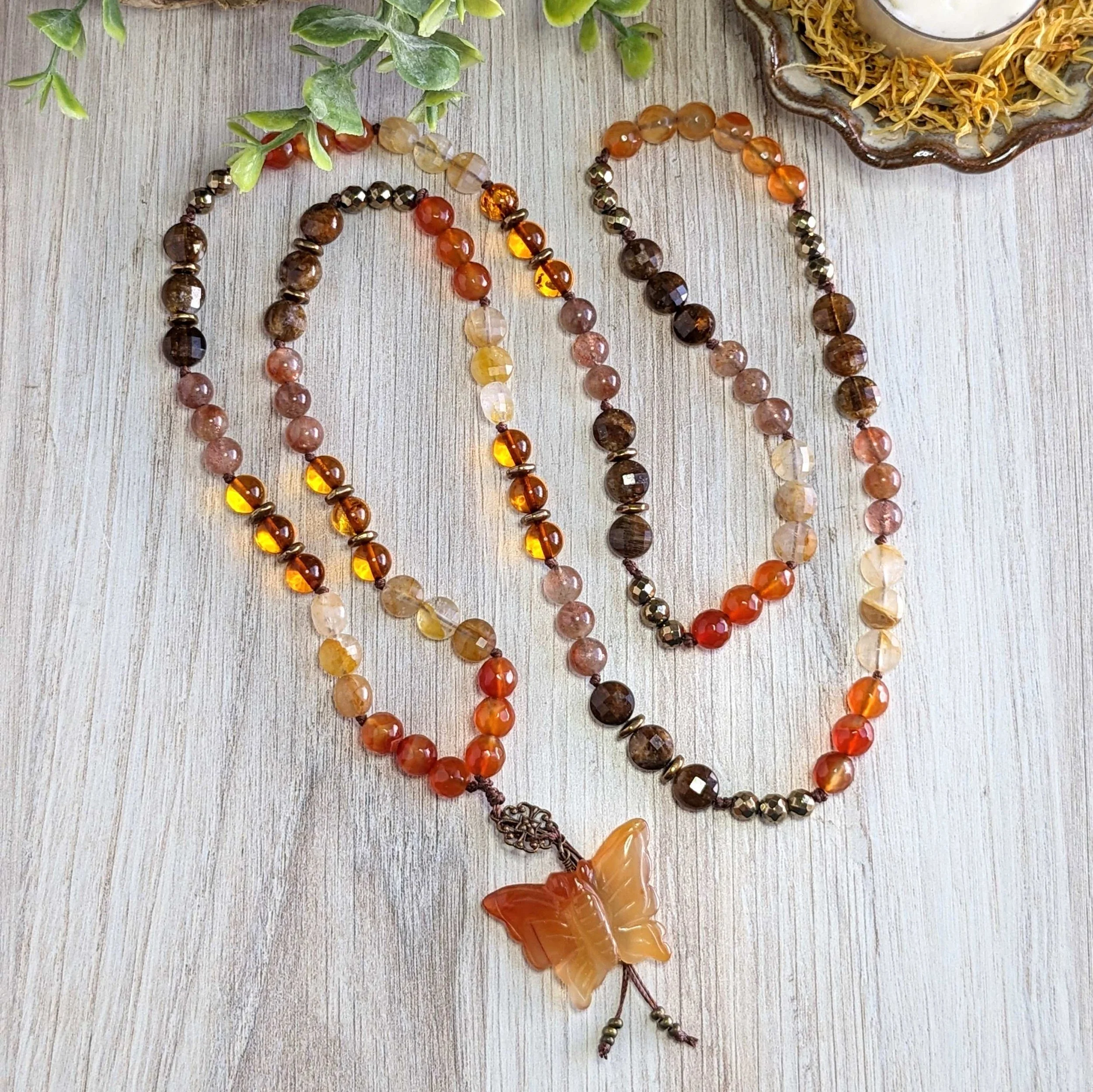 Three beaded necklaces with amber colors, featuring a butterfly-shaped pendant, are laid out on a wooden surface with some green foliage and a decorative dish in the background.