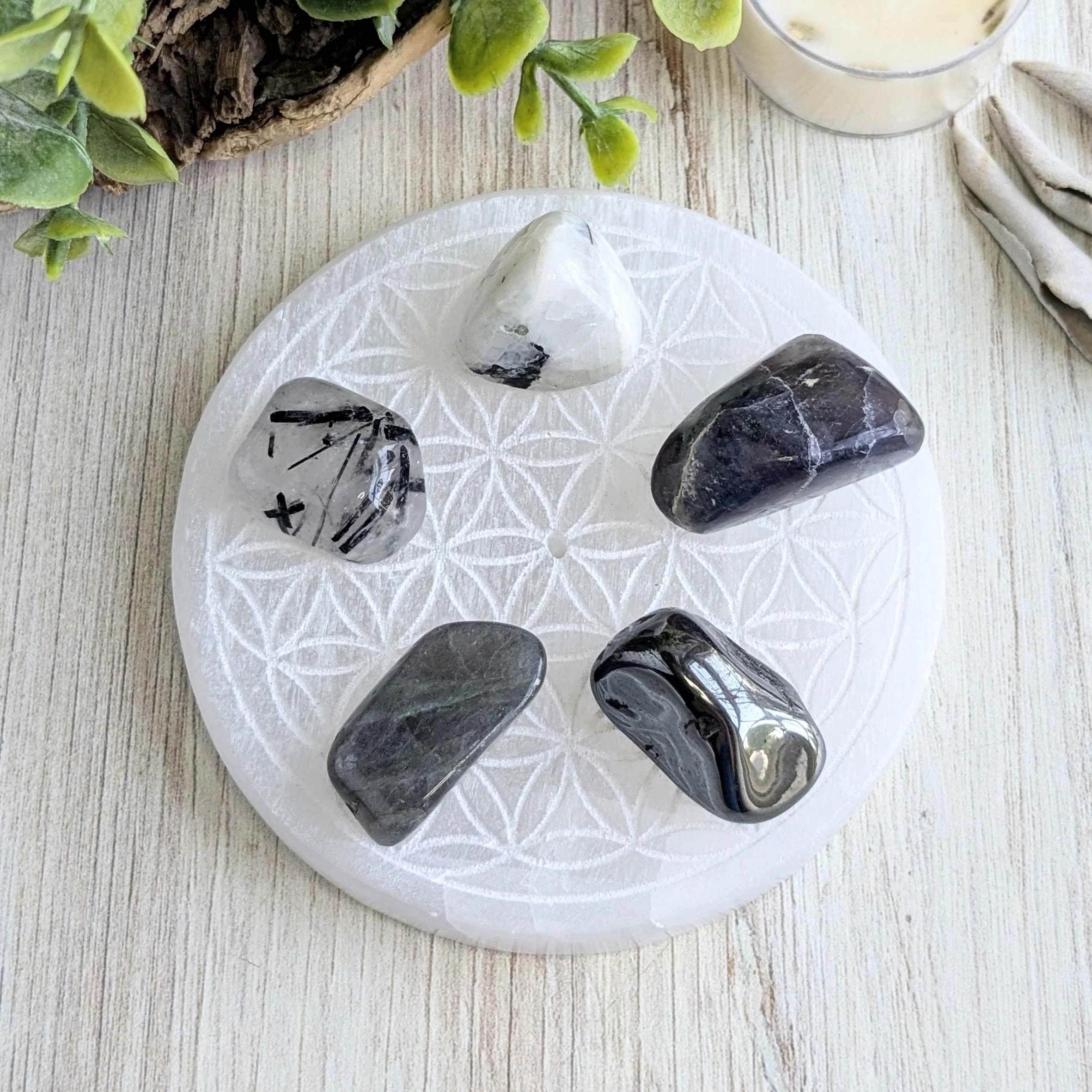 Five polished black and white stones arranged on Selenite on a decorative white plate, with a plant, a candle, and a cloth nearby on a wooden surface.