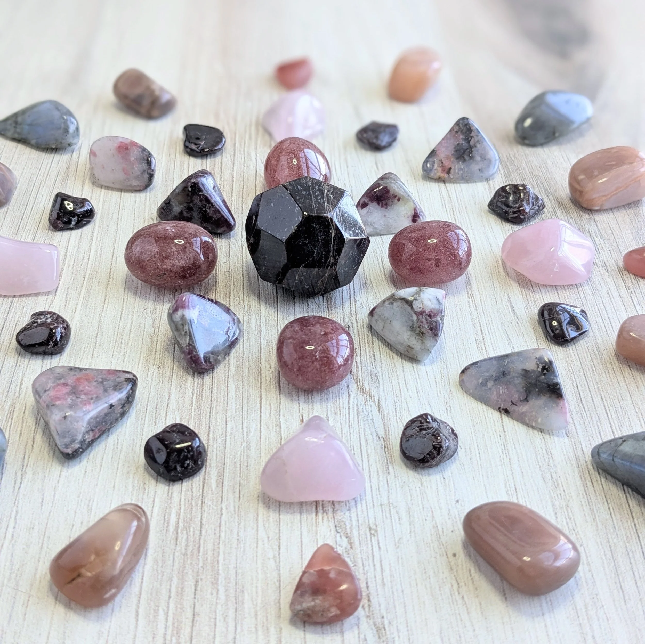 Various polished gemstones surrounding an Almandine Garnet, pink, red, and black stones, scattered on a light wooden surface.