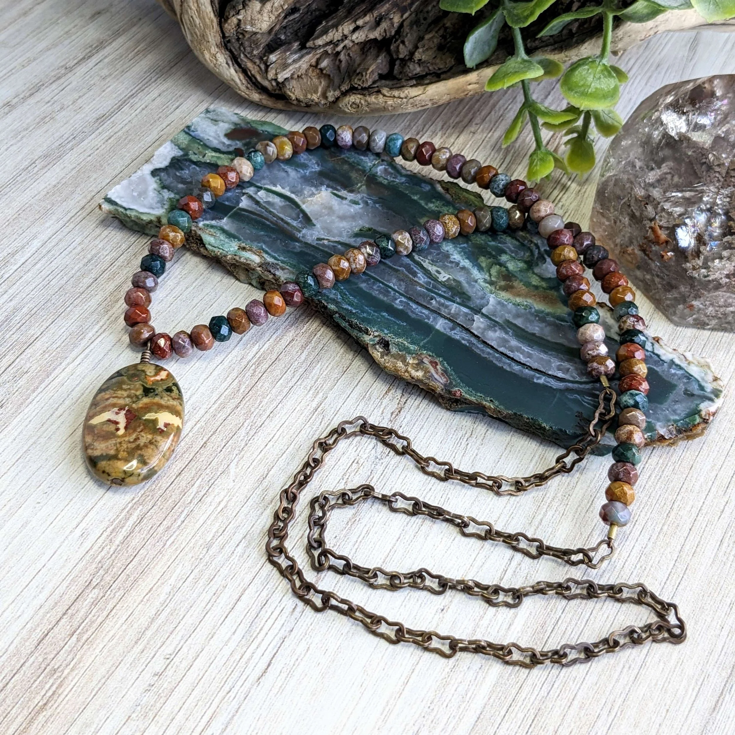 Colorful beaded necklace with a large oval pendant, resting on a polished stone slab, with green plant and crystal decor in the background.