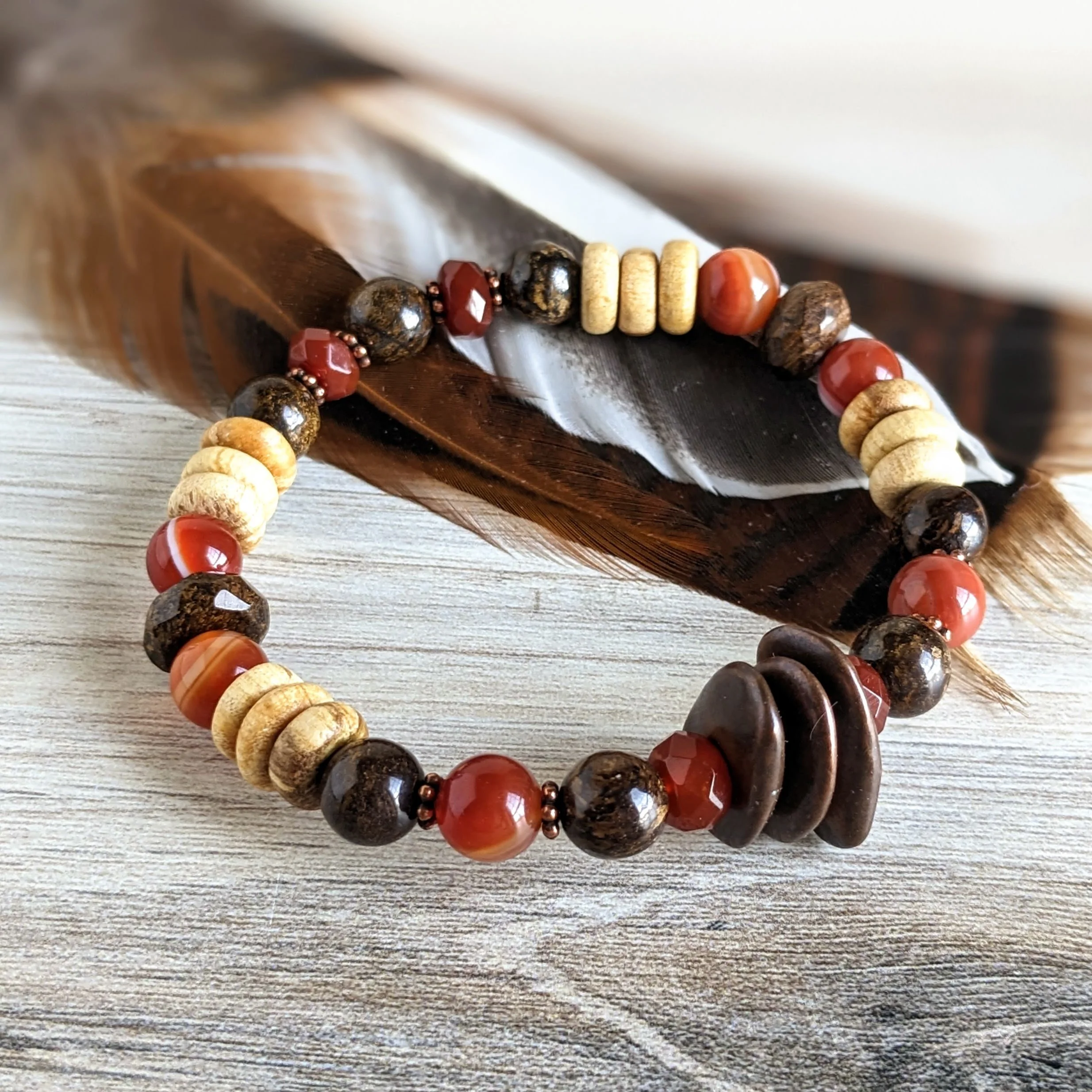 Beaded bracelet with brown, red, and beige stones resting on a feather and wooden surface.