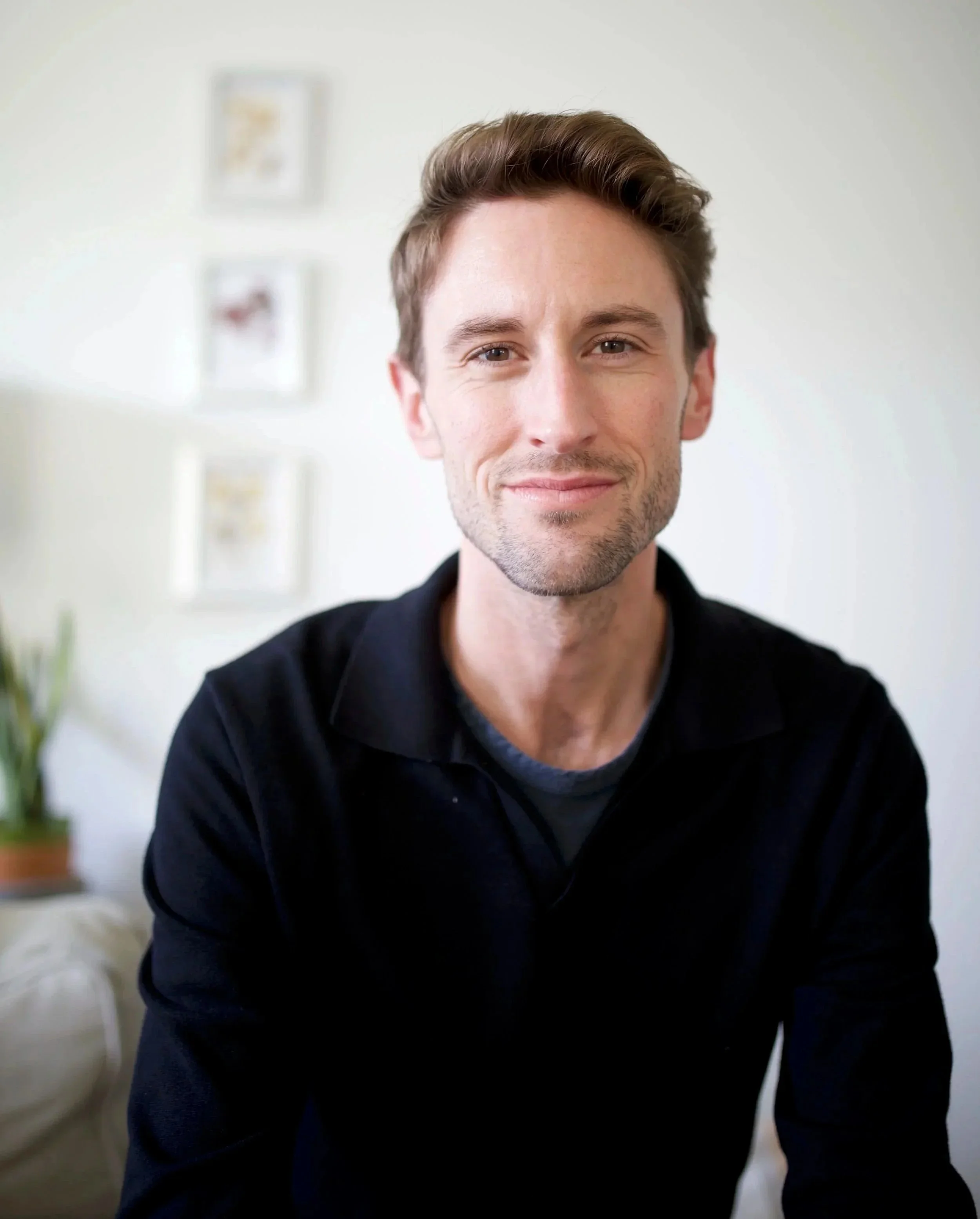 A smiling man with light brown hair and stubble, wearing a black jacket, in a well-lit room with a white wall and blurred framed pictures in the background.