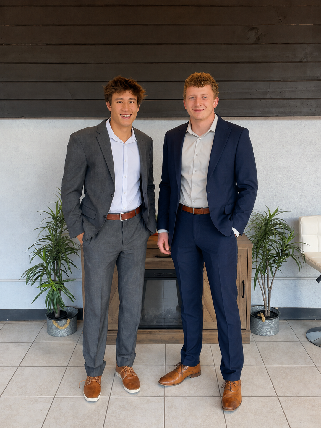 Two young men in business suits standing indoors, smiling for the camera. They are in front of a wooden cabinet with plants on either side.