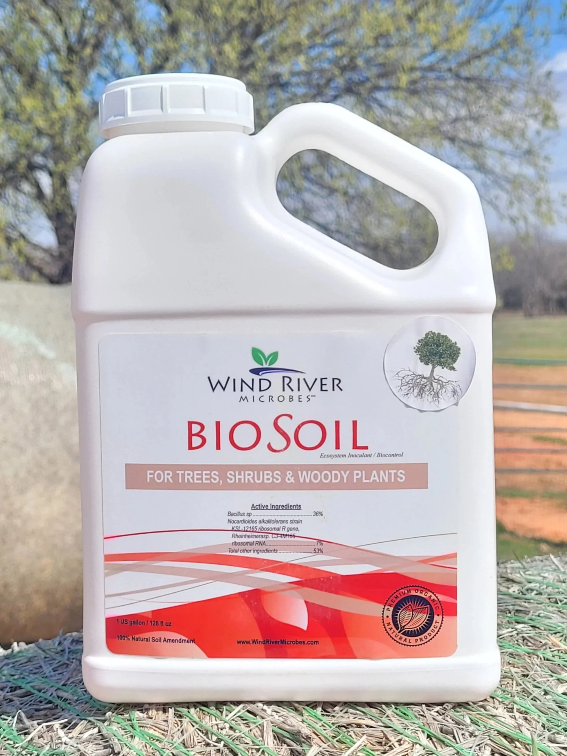 A large white container of Wind River Microbes BioSoil for trees, shrubs, and woody plants, placed outdoors on a textured surface with a tree and a grassy field in the background.