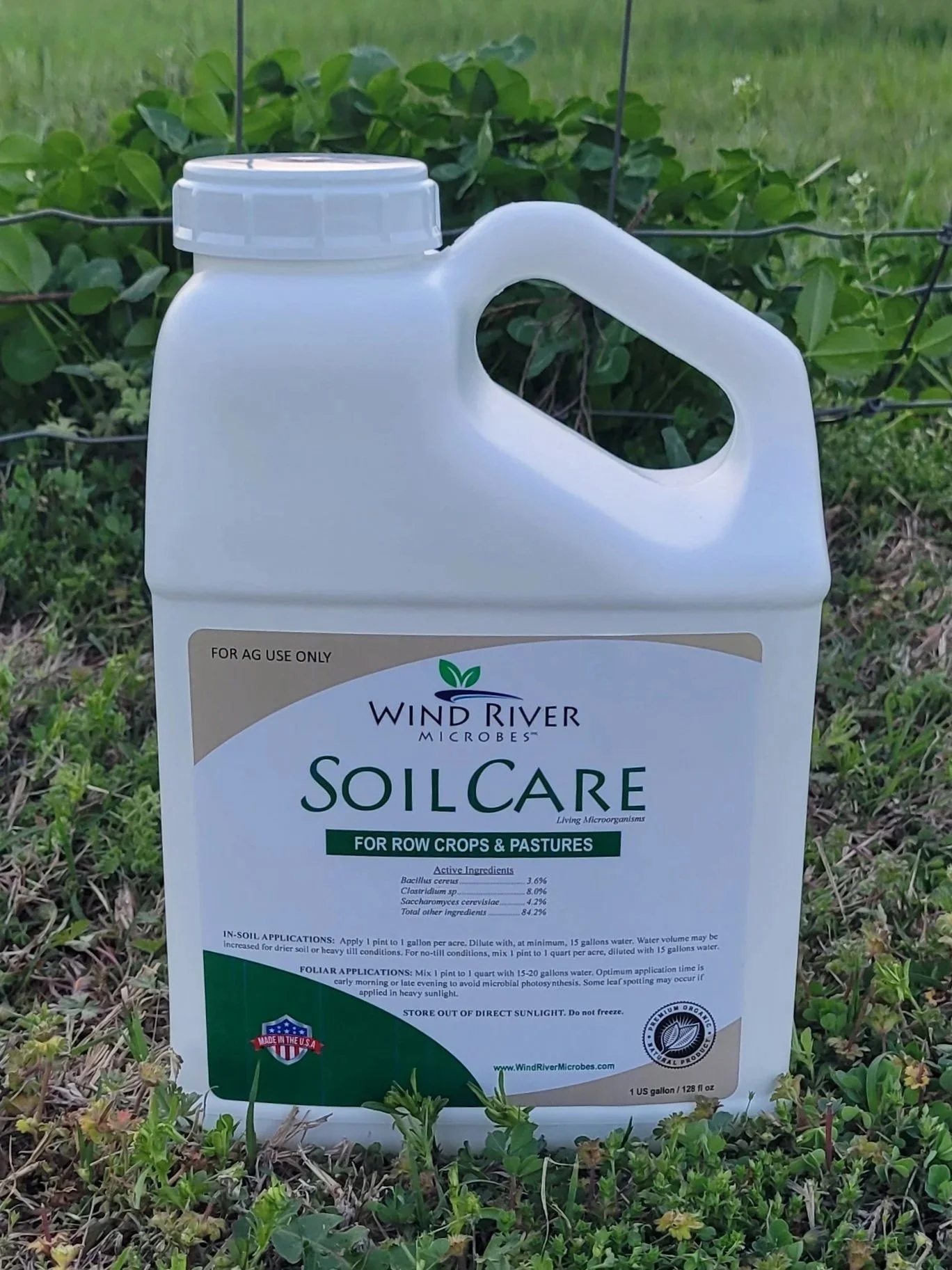 A large white container of SoilCare by Wind River Microbes, designed for row crops and pastures, placed outdoors on grass with a black wire fence and green plants in the background.