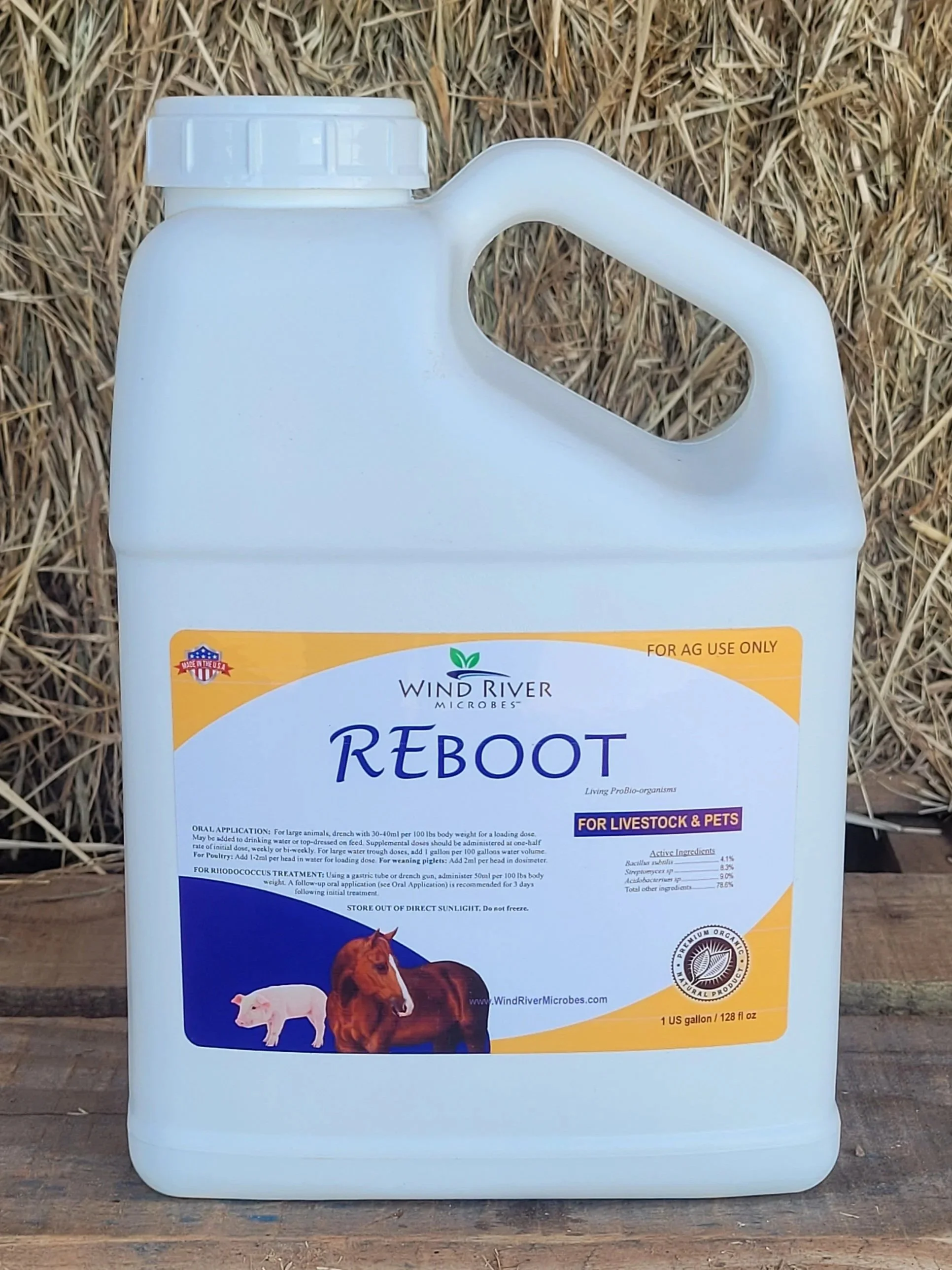 White 1-gallon container of Wind River Microbes Reboot, a probiotic supplement for livestock and pets, placed on a wooden surface with hay in the background.