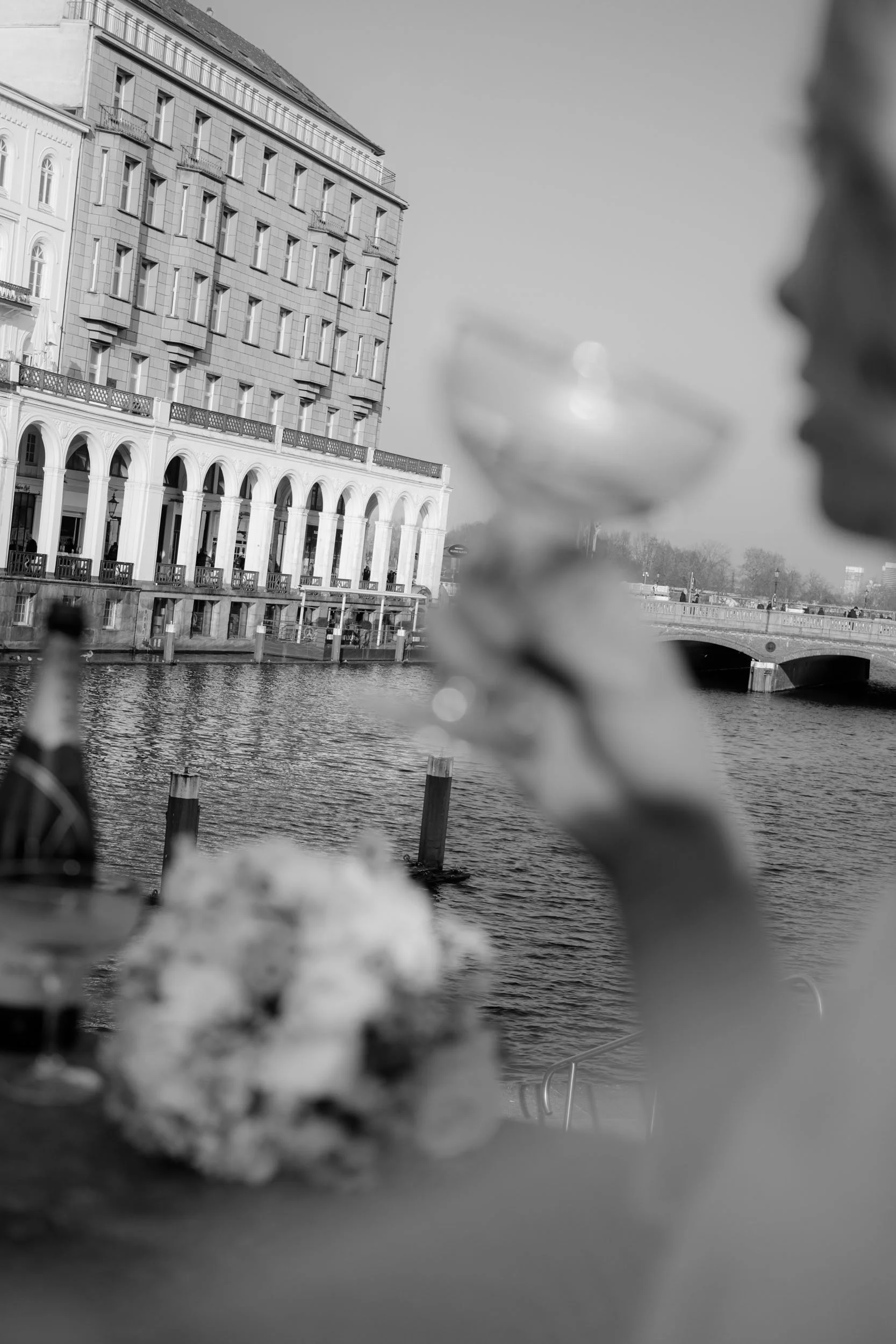 Editorial Braut Shooting an den Alsterarkaden in Hamburg: Eine Braut (unscharf im Vordergrund) stößt mit Champagner vor der historischen Binnenalster an. Zeitlose Hochzeitsfotografie von Valeska Weddings.