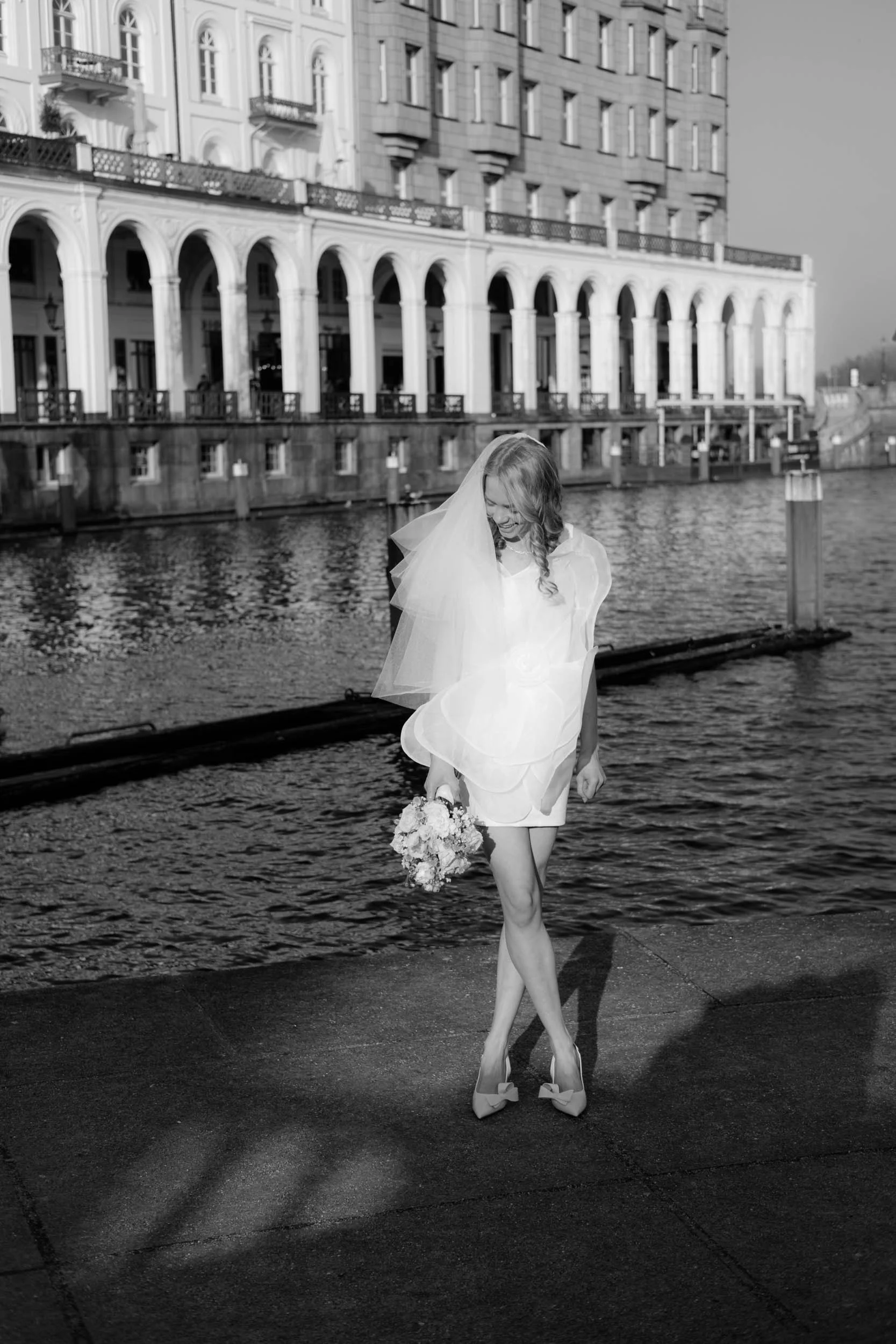 Editorial Brautshooting an den Alsterarkaden in Hamburg: eine lachende Braut in einem kurzen modernen Hochzeitskleid vor historischer Kulisse eingefangen von Hochzeitsfotografin Valeska Weddings.