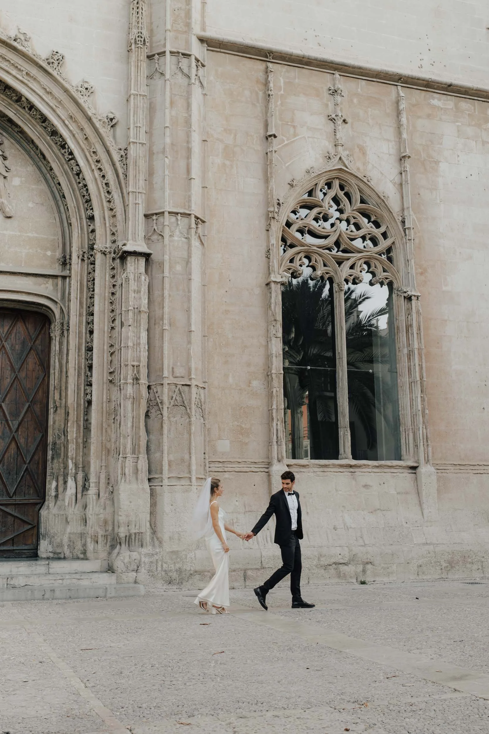 Editorial Destination Wedding in Palma de Mallorca: Ein Brautpaar läuft Hand in Hand vor der beeindruckenden gotischen Architektur der Altstadt Palma. Authentisches Storytelling von Valeska Weddings.