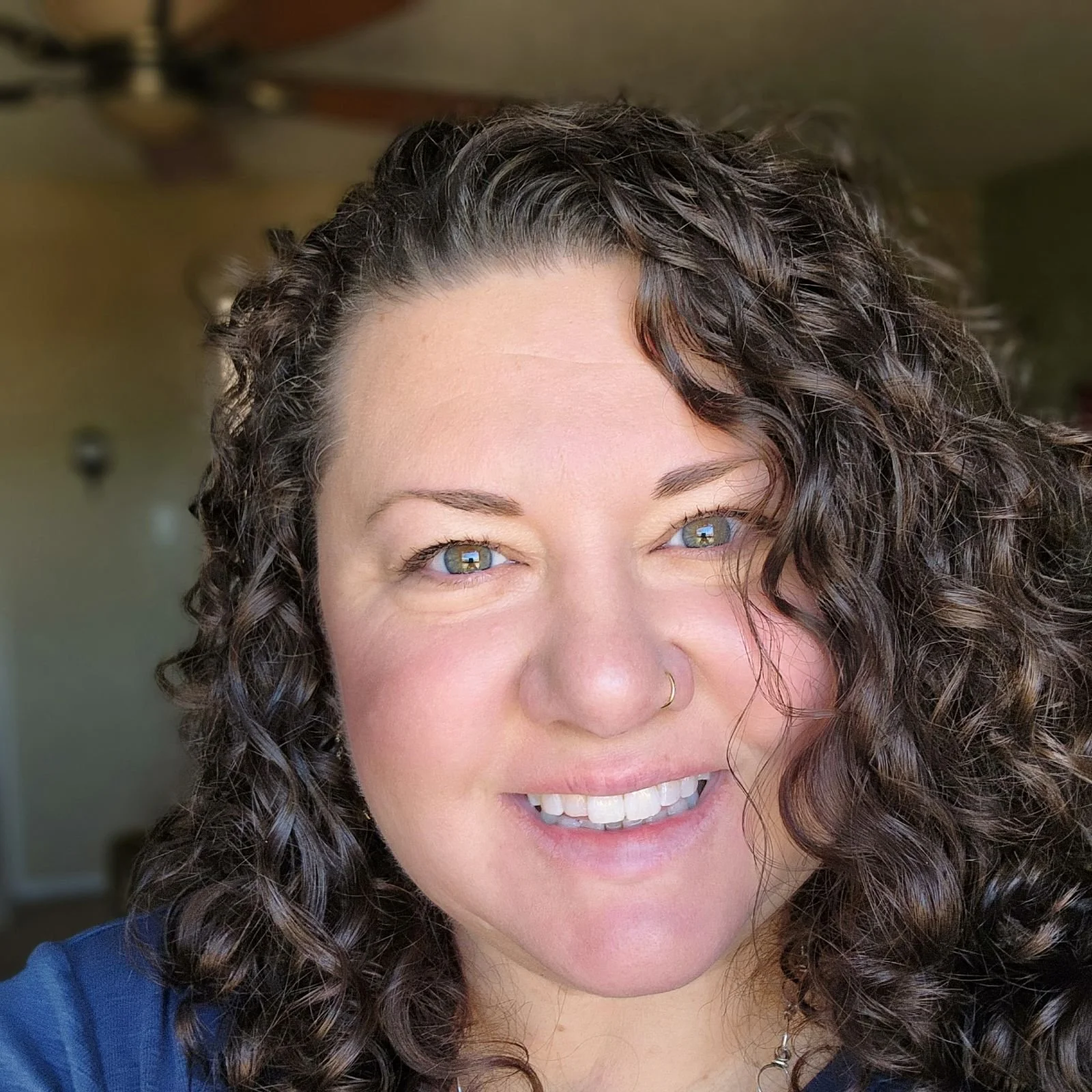 Close-up of a smiling woman with curly dark brown hair, blue eyes, light makeup, and a nose piercing.