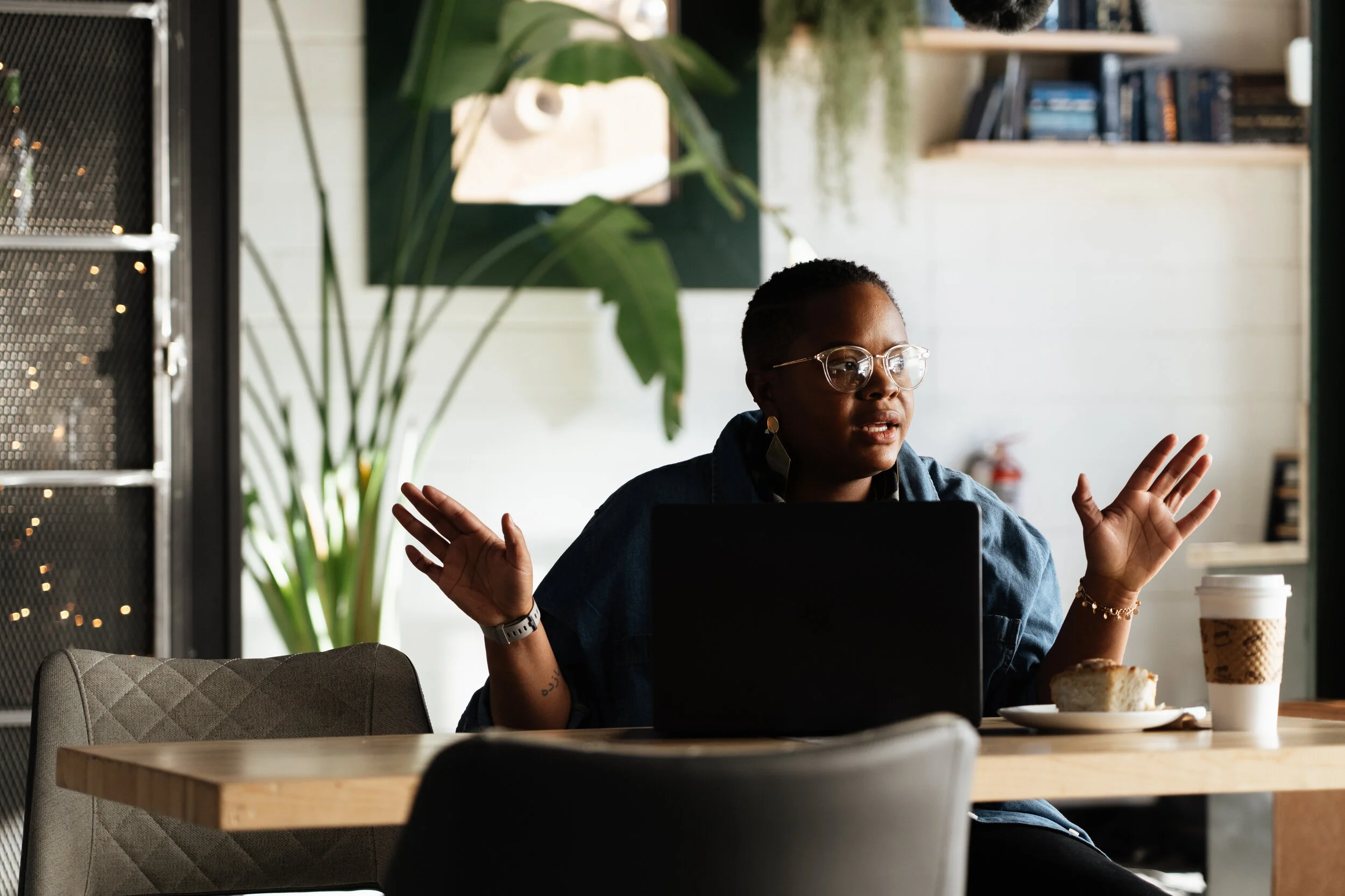 A woman with dark skin, wearing glasses, sitting at a wooden table in a cafe or workspace, with a laptop, a plate with a cake, and a to-go coffee cup, gesturing with her hands while talking or explaining.