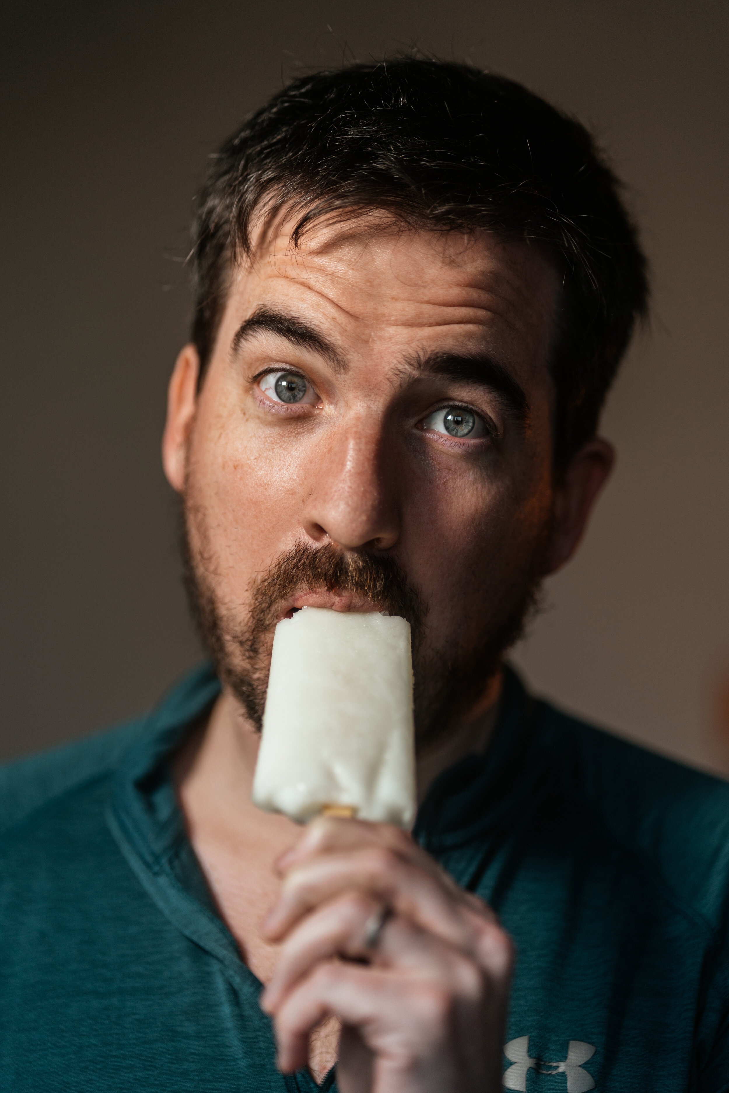 A man with dark hair and a beard, wearing a teal hoodie, is licking a white popsicle.