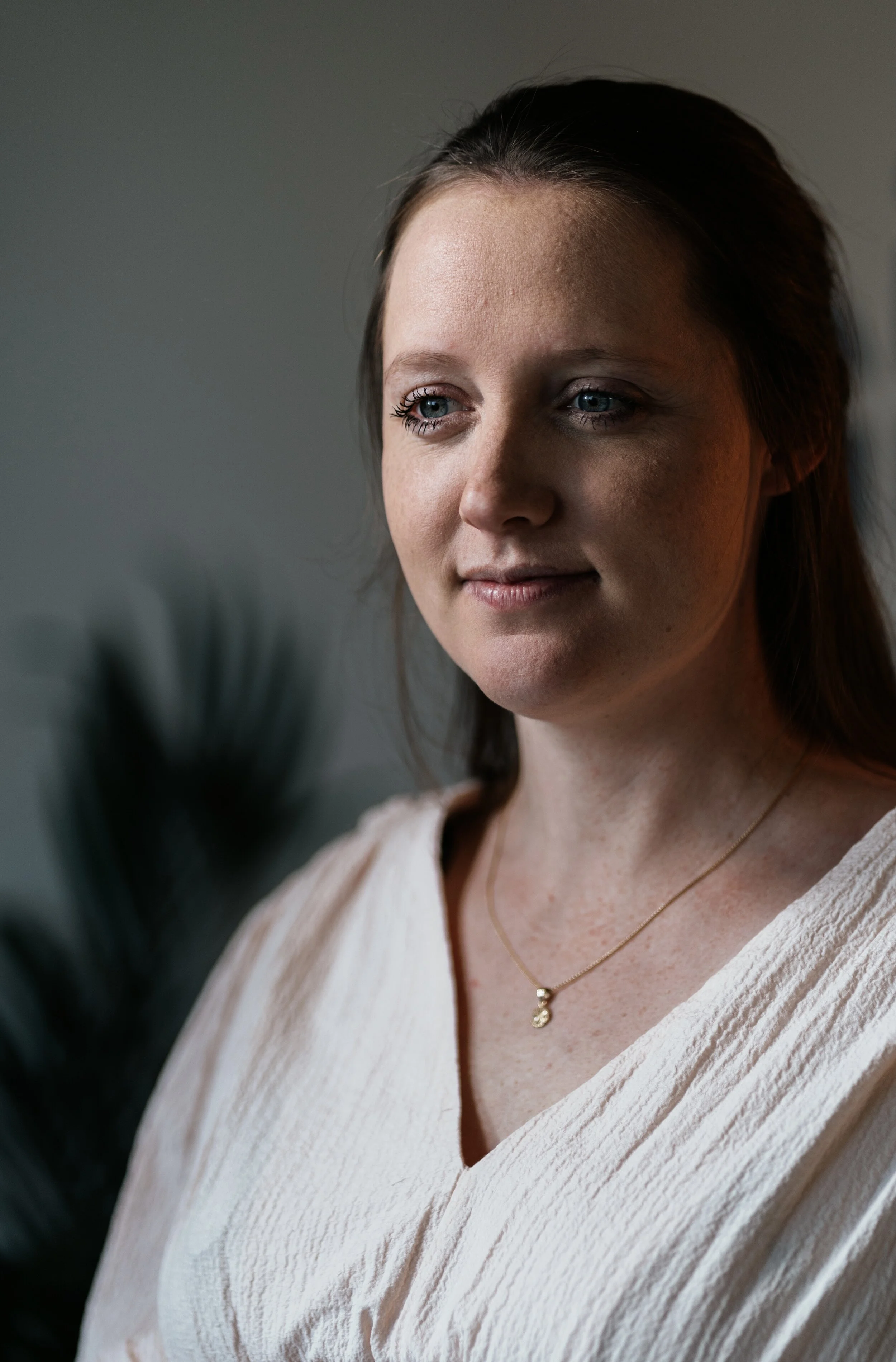 A woman with light skin and brown hair, wearing a white textured blouse and a delicate gold necklace with a small pendant, gazing downward in a softly lit indoor setting.