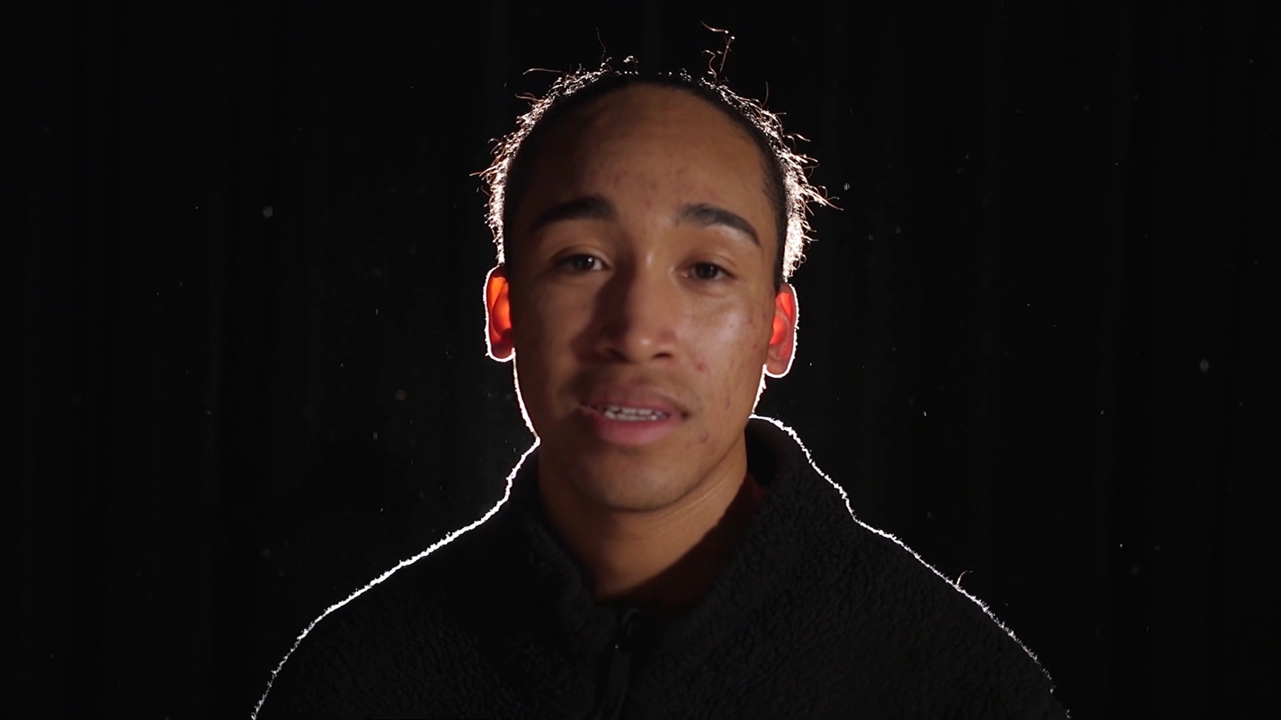 Close-up of a young man with dark hair, wearing a black fuzzy jacket, standing against a black background illuminated by backlighting that outlines his face and shoulders.