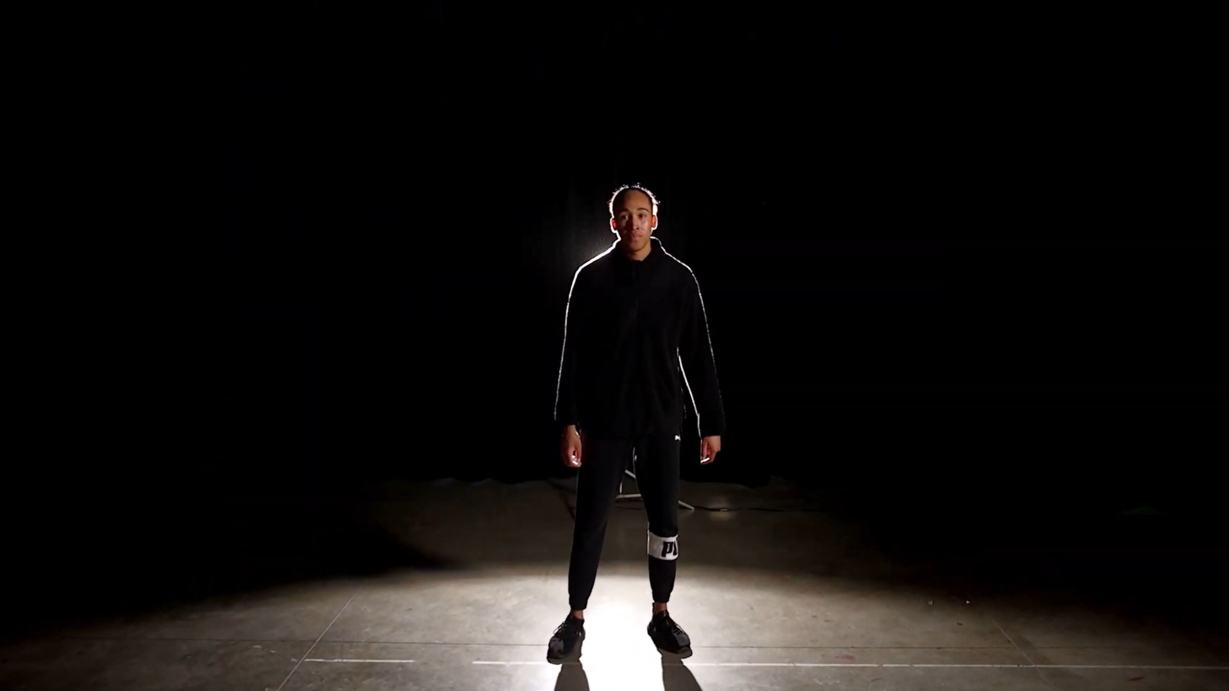 A young man stands in the center of a dimly lit room, illuminated from behind creating a glow around him, with a dark background and visible floor tiles.