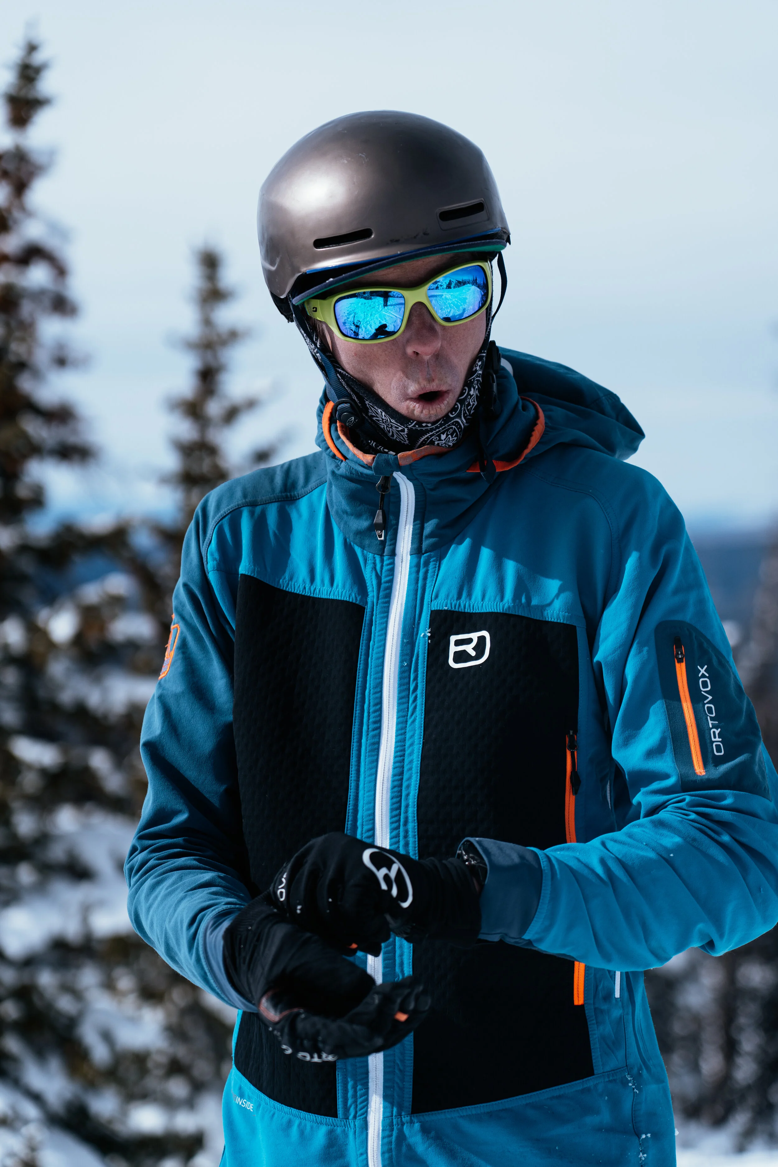A man wearing a helmet, reflective sunglasses, and a blue jacket with black and orange accents outdoors in a winter setting with snow and trees.